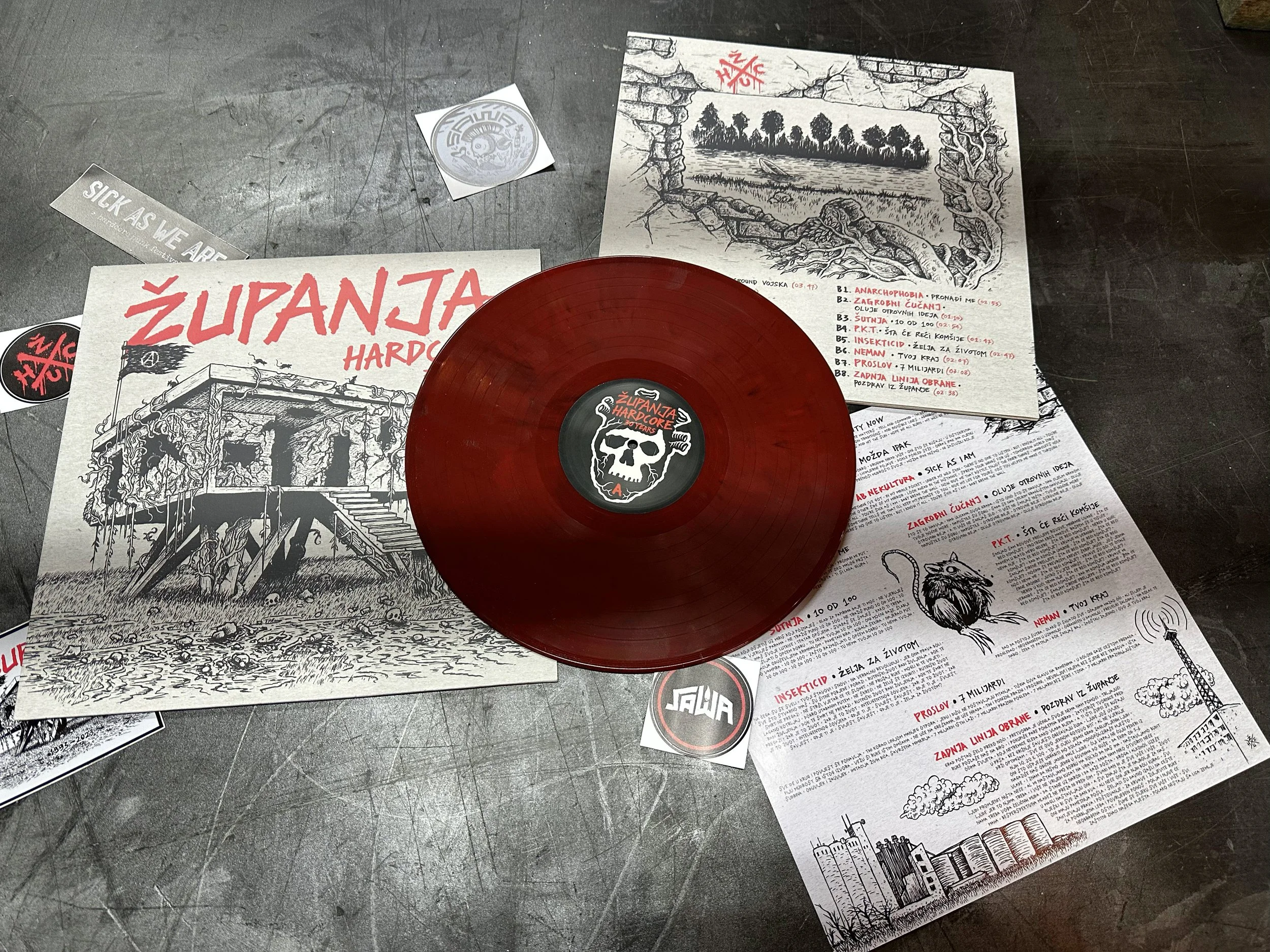 A red vinyl record with a sticker featuring a skull and a black and white coloring book page with a detailed drawing of a ruined structure and a lake in the background, along with various paper sheets and stickers on a dark surface.