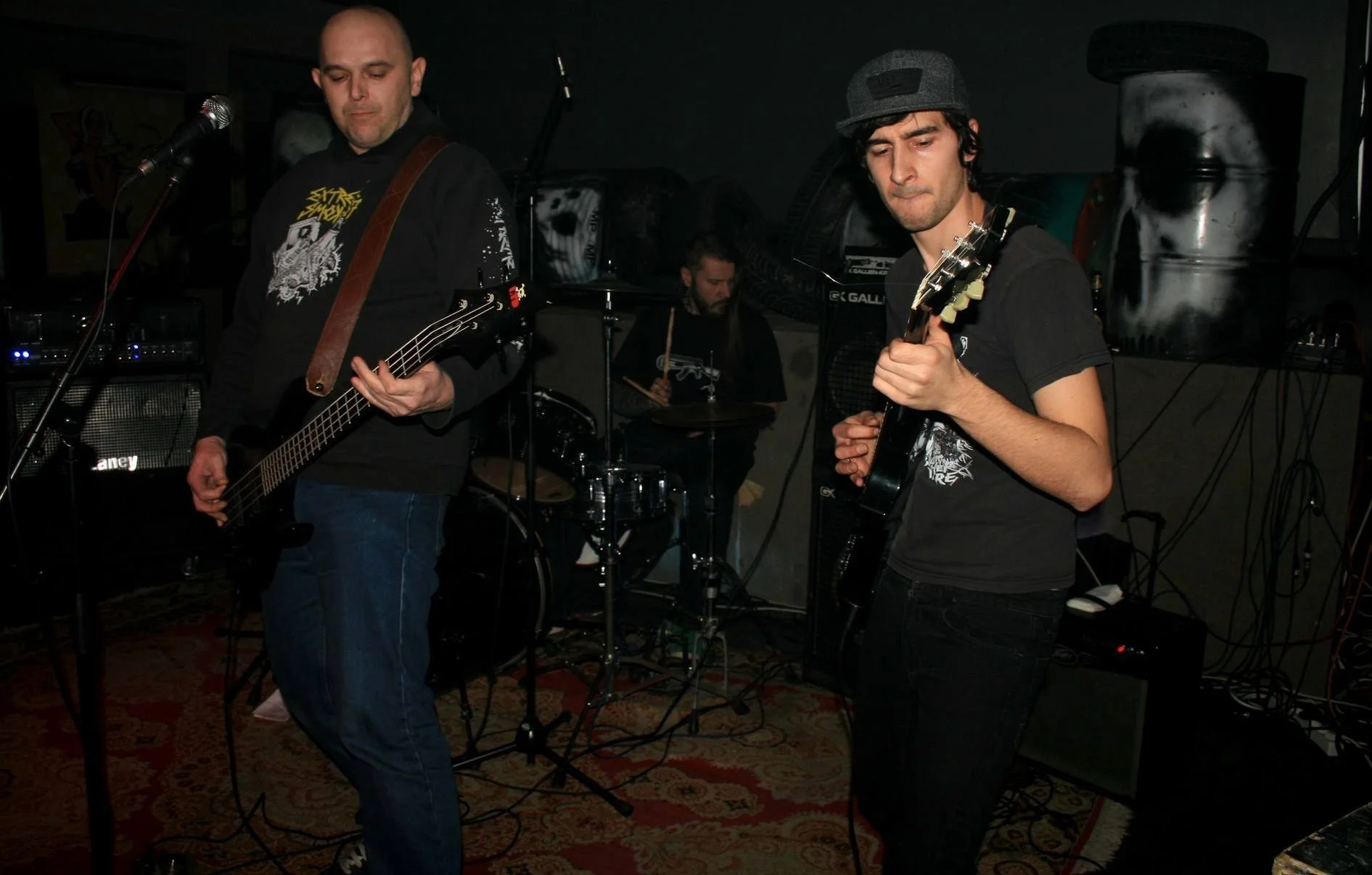Three musicians performing in a dark, intimate venue. Two guitarists are in the foreground, one with a bass guitar, and a drummer in the background playing a drum set. The guitarists are focused on their instruments, with the one on the right wearing