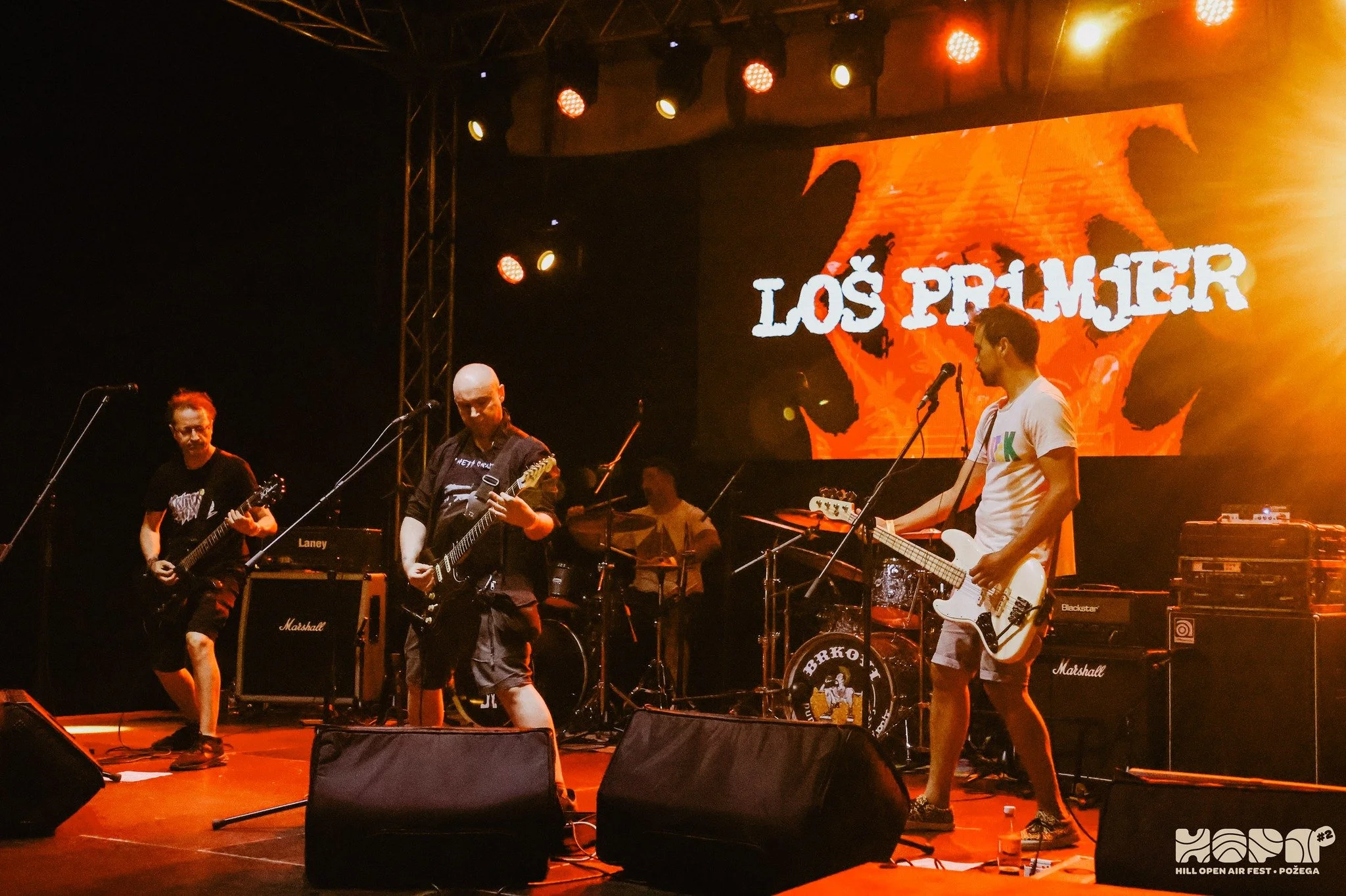 A band performing on stage at Hill Open Air Fest in Požega, with musicians playing guitars and drums, and a backdrop displaying 'Los Primer' with a fiery background.