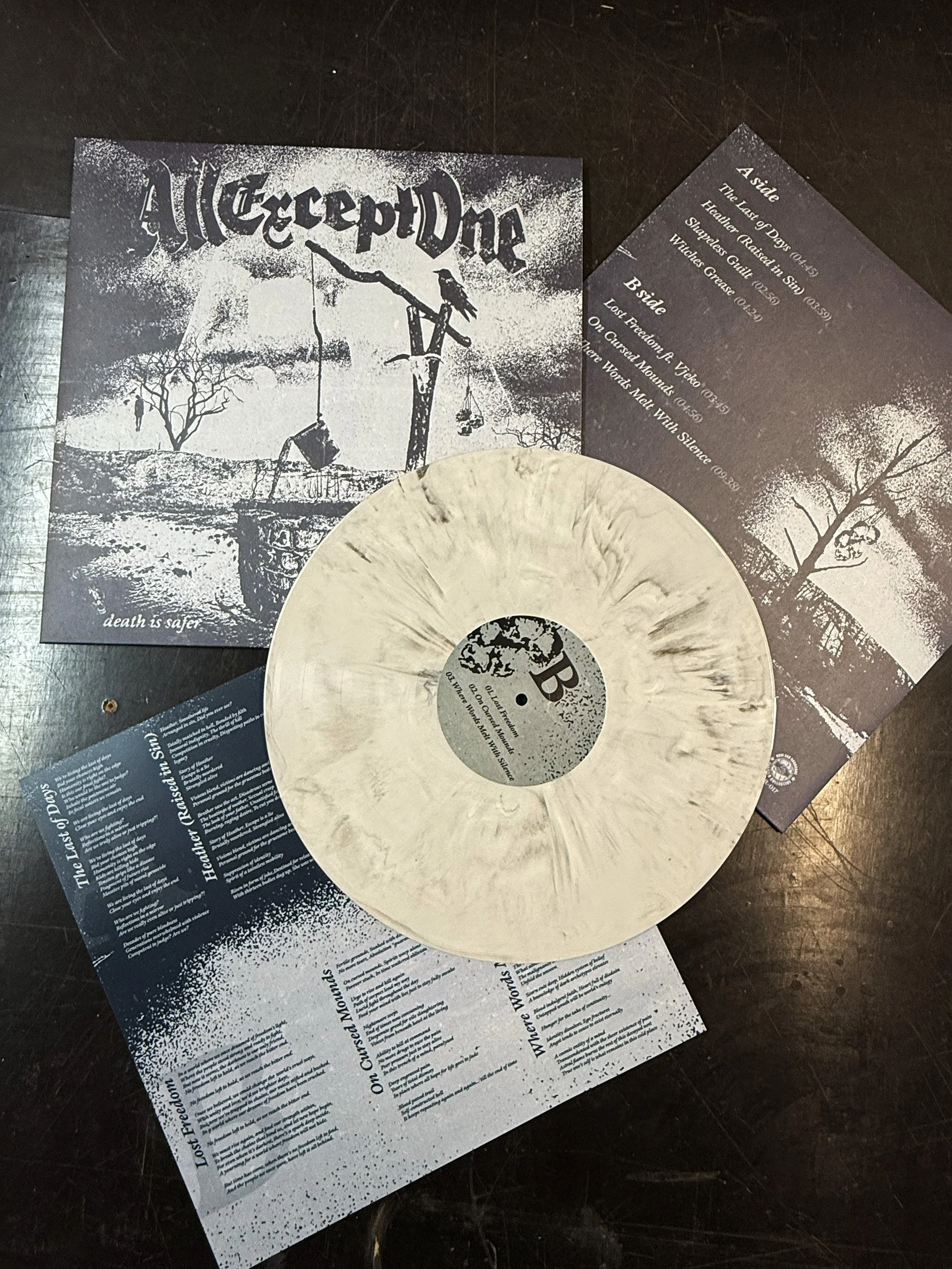 A black and white record with a splattered design, placed on top of a black surface. Behind it, there are several printed pages or flyers with text about a music album or event, featuring themes of death, freedom, and darkness.