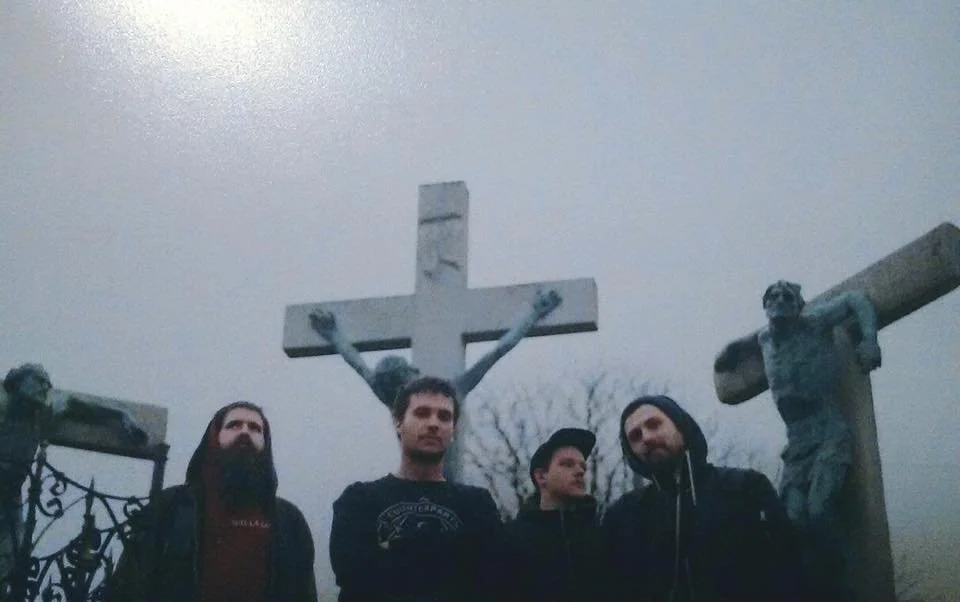 Four men standing in front of three crosses in an outdoor setting with a gray sky.