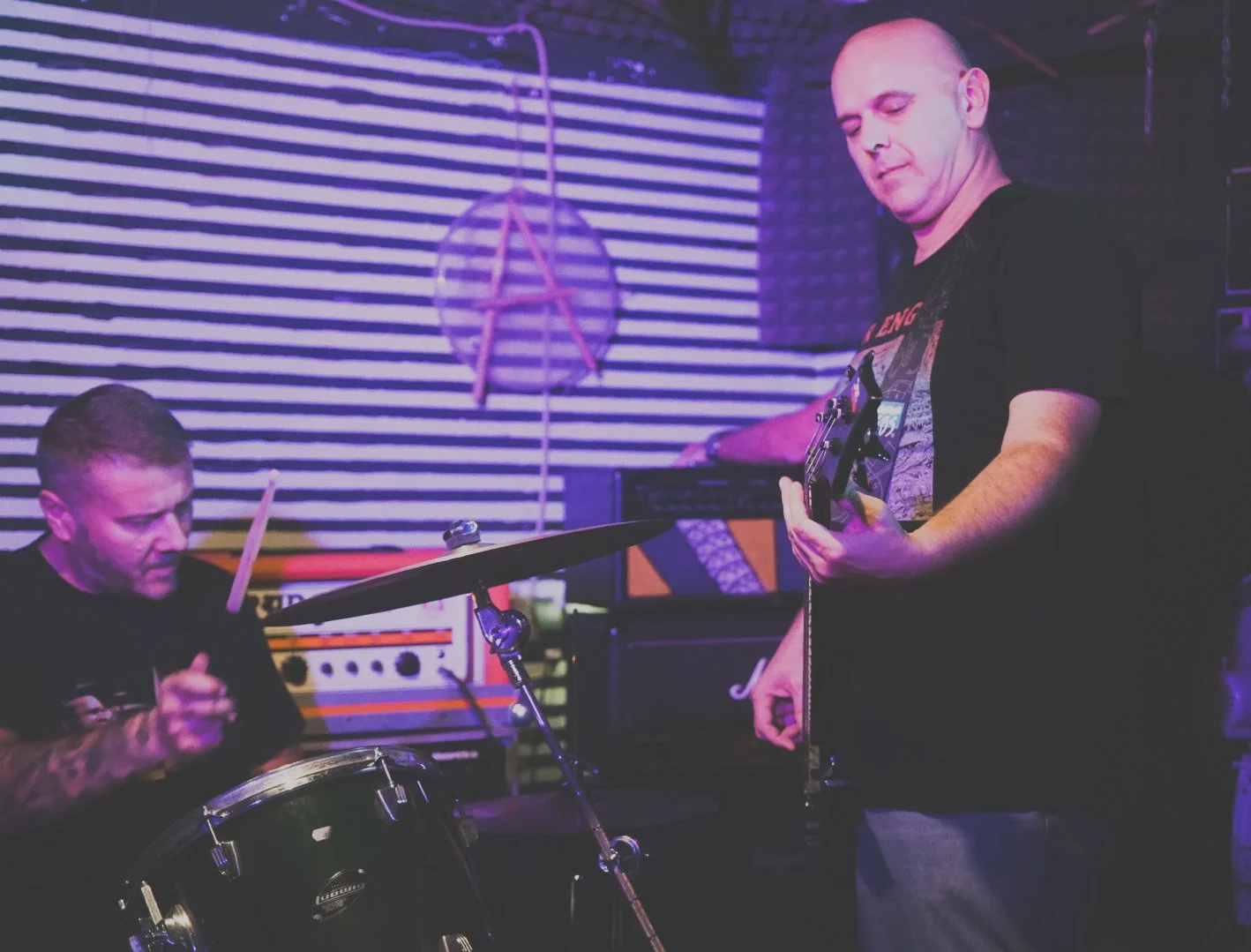 Two men playing musical instruments in a music studio with purple and black decor. One is playing drums and the other is playing an electric guitar.