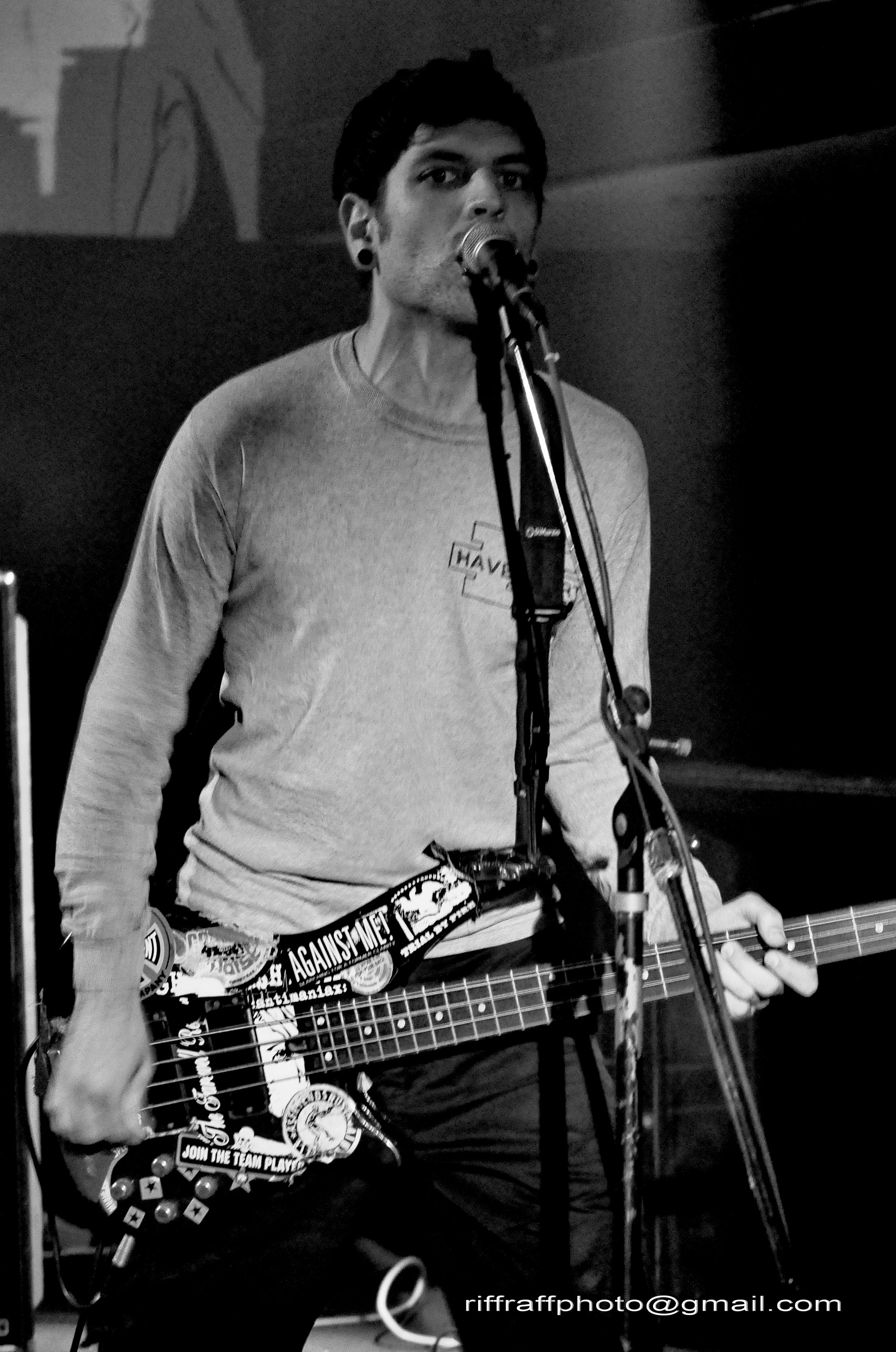 Black and white photo of a person singing into a microphone while playing an acoustic guitar, wearing a light-colored shirt with patches and stickers, and standing in front of a dark background.