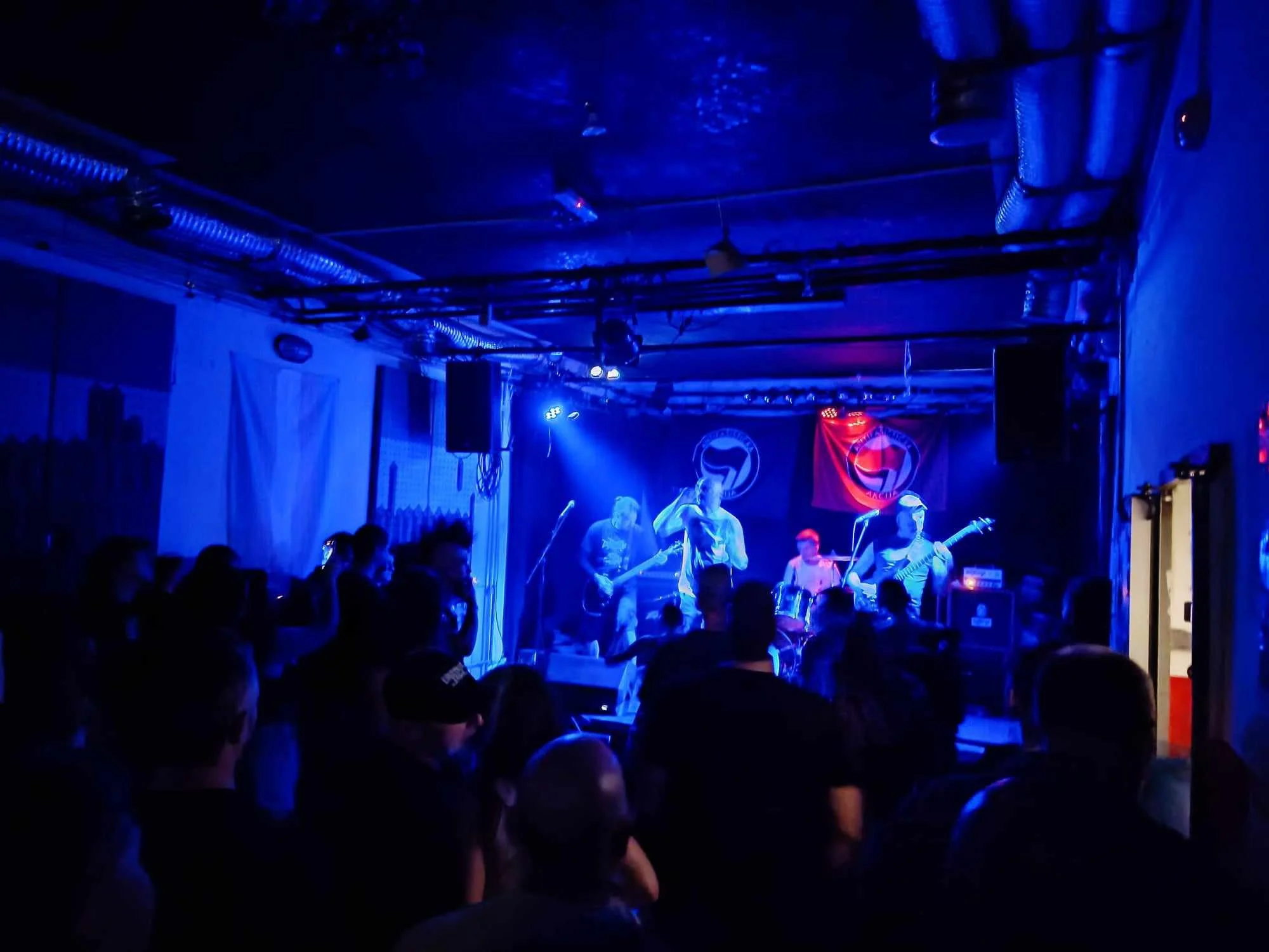 A live music performance in a dimly lit venue with blue lighting, showing a band on stage with members playing guitars and singing, and an audience watching.