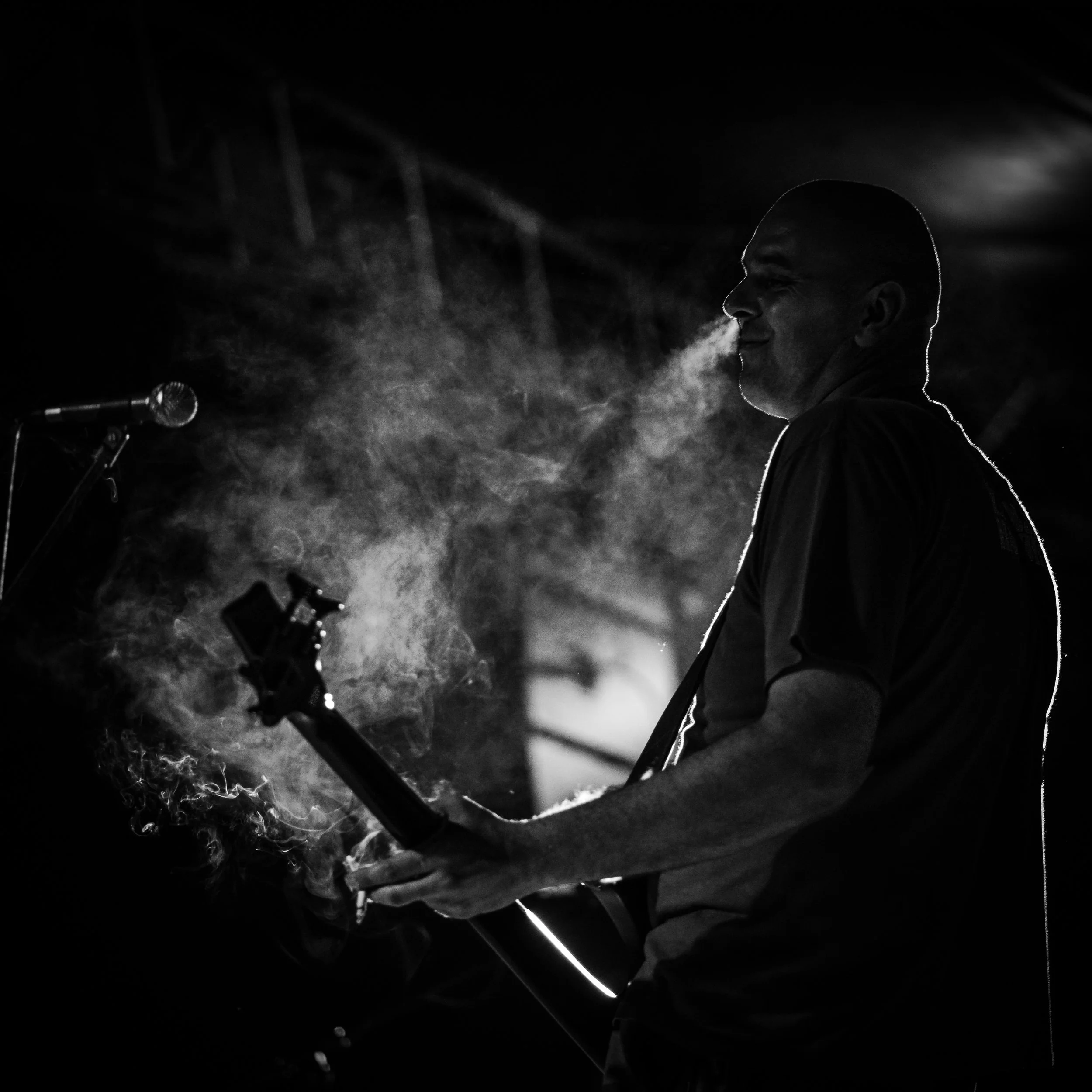 Silhouette of a man with a shaved head, smiling and exhaling vapor or breath in a dark setting, with a microphone and stage equipment visible.
