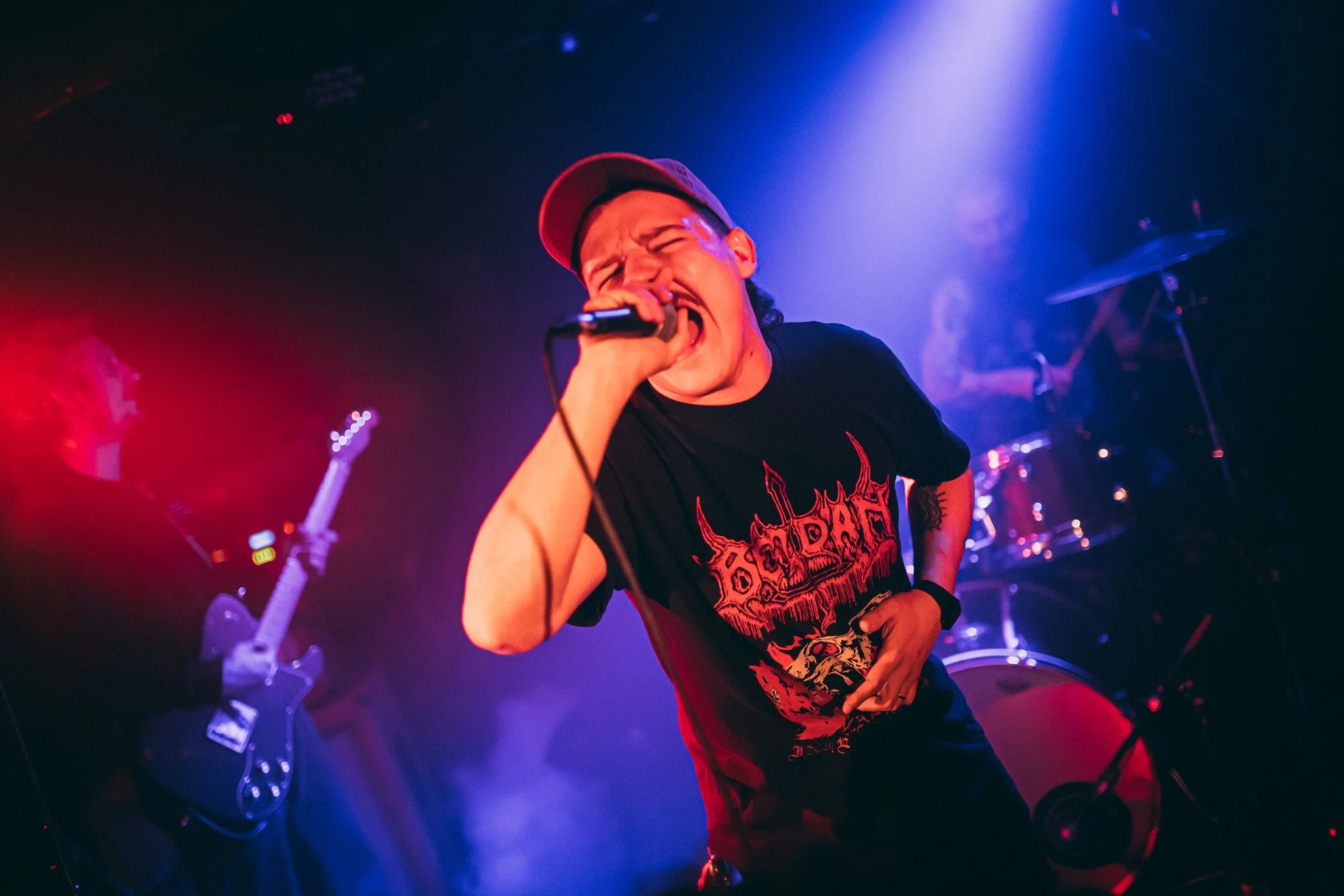 A man wearing a red hat and a black t-shirt with a graphic design is singing into a microphone on stage under colorful stage lights. There is a guitar and drums in the background.