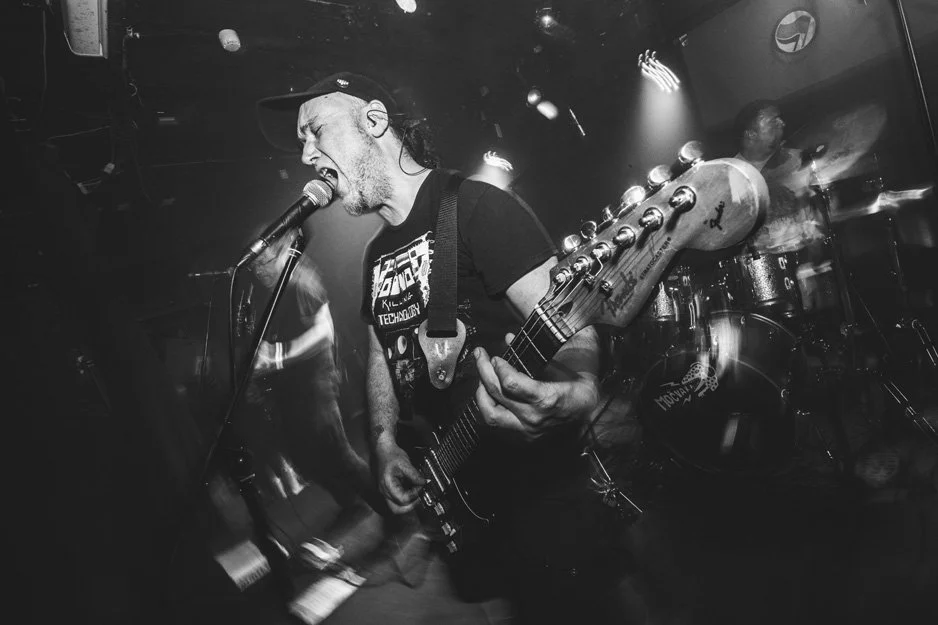 A musician passionately singing into a microphone while playing an electric guitar on stage, with a drummer in the background.