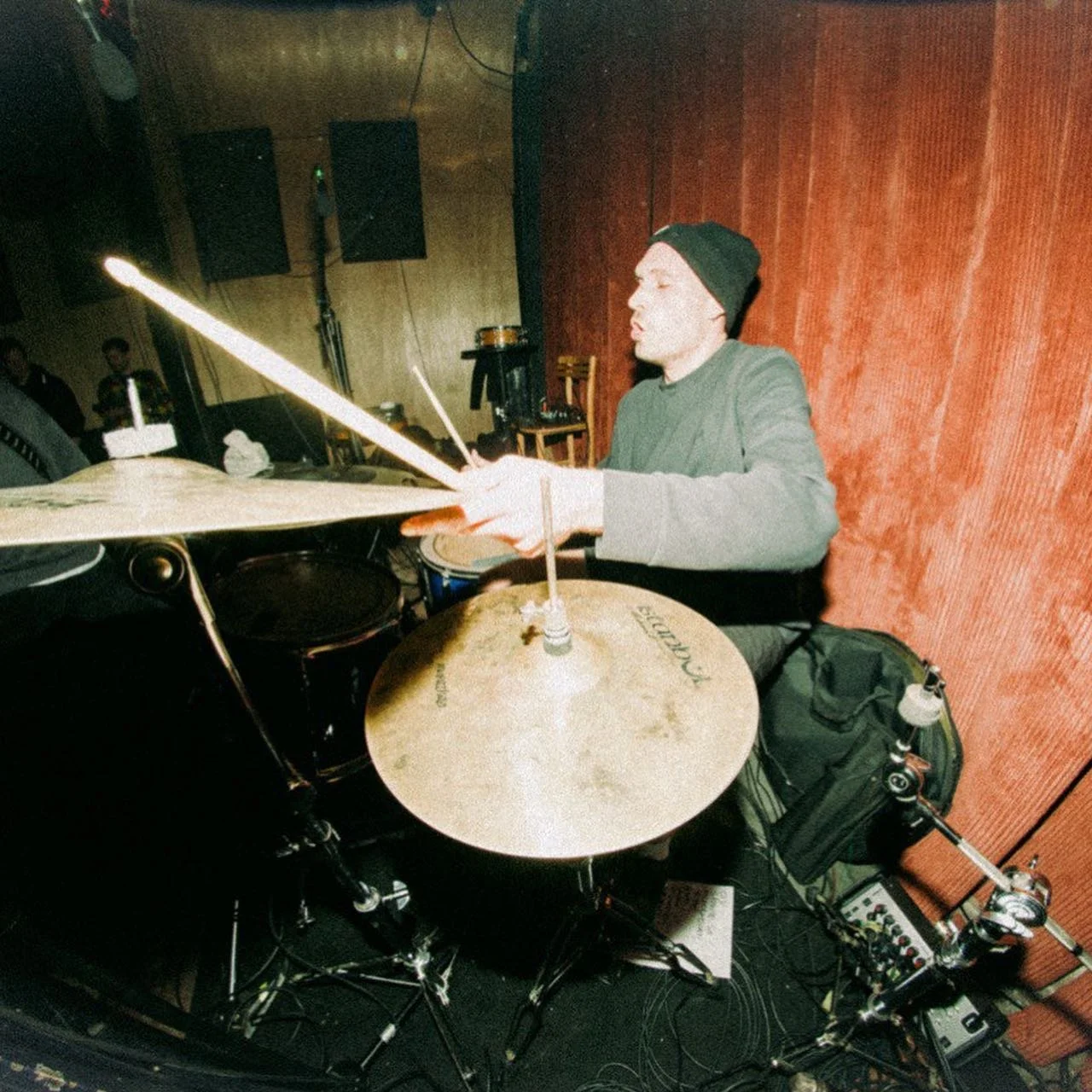 A man wearing a black beanie playing a drum set in a room with red and wooden walls.