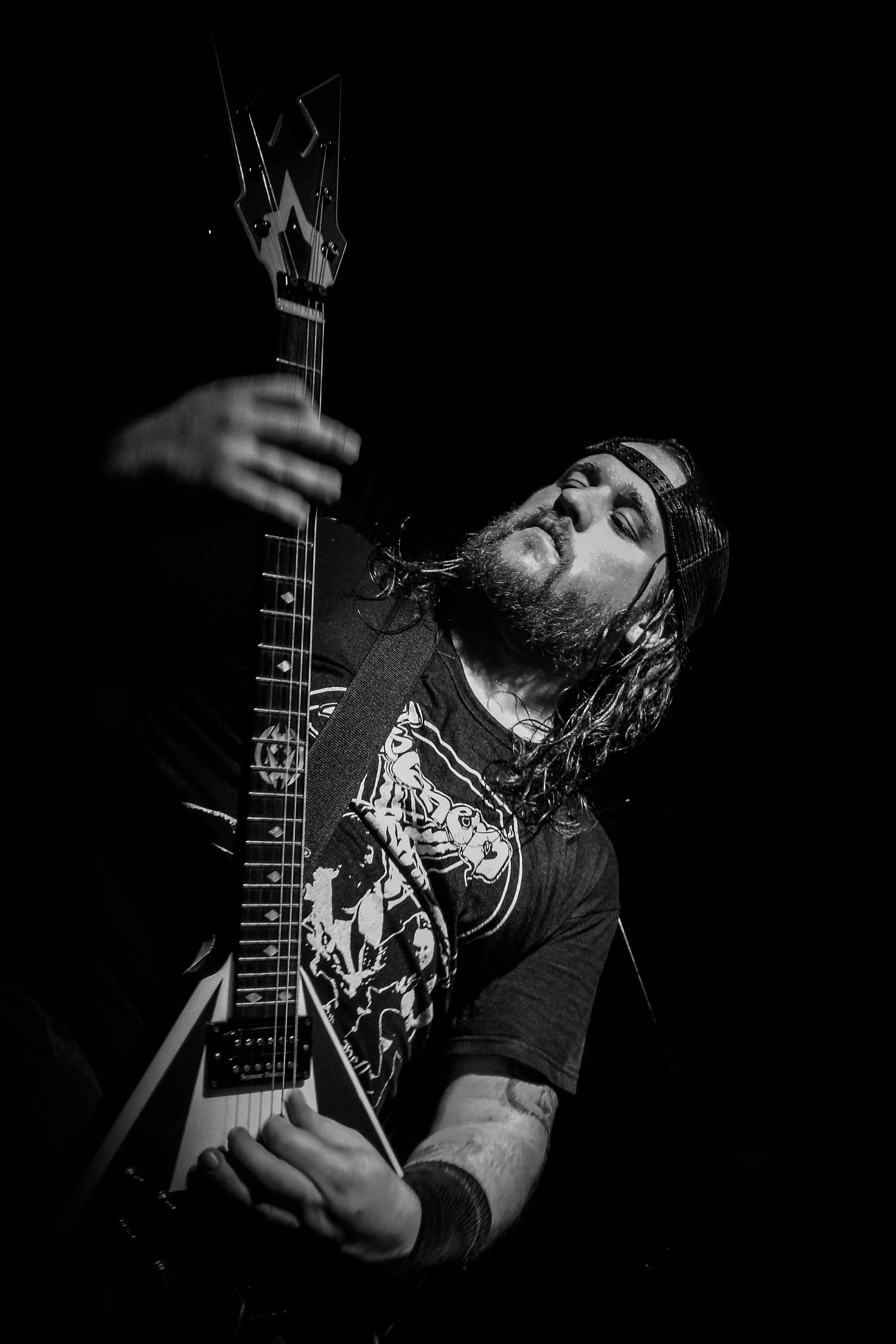 A musician with long hair and a beard is playing an acoustic guitar on stage. He is wearing a backwards cap and a graphic T-shirt, and appears to be singing or passionately performing.