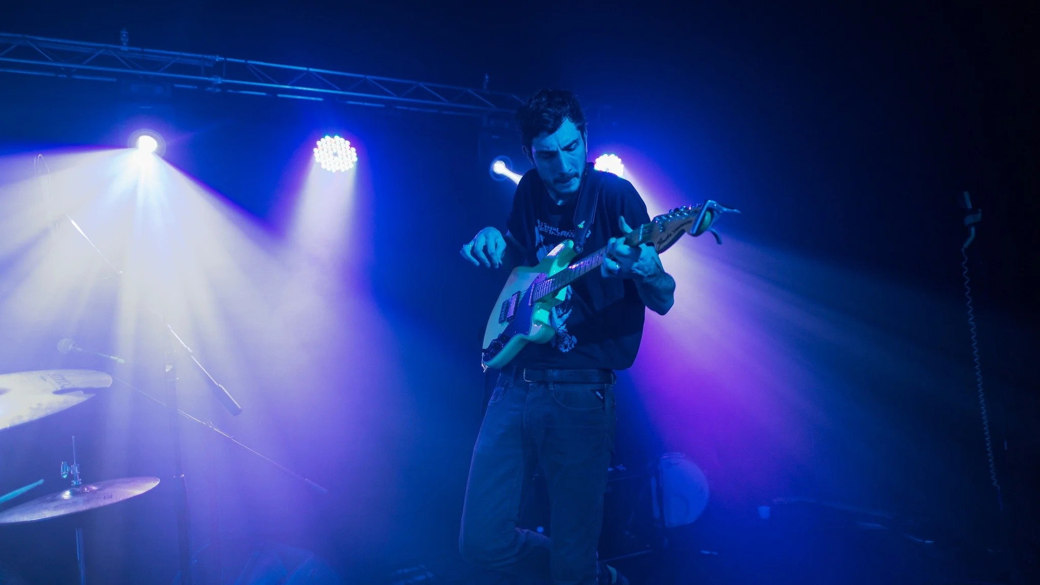 Male musician playing an electric guitar on stage under blue and purple lighting.