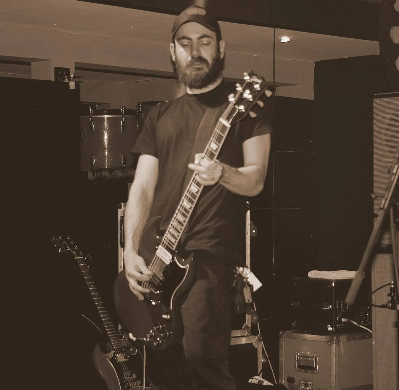 A man with a beard and cap playing an electric guitar on stage, with drums and other musical equipment in the background.