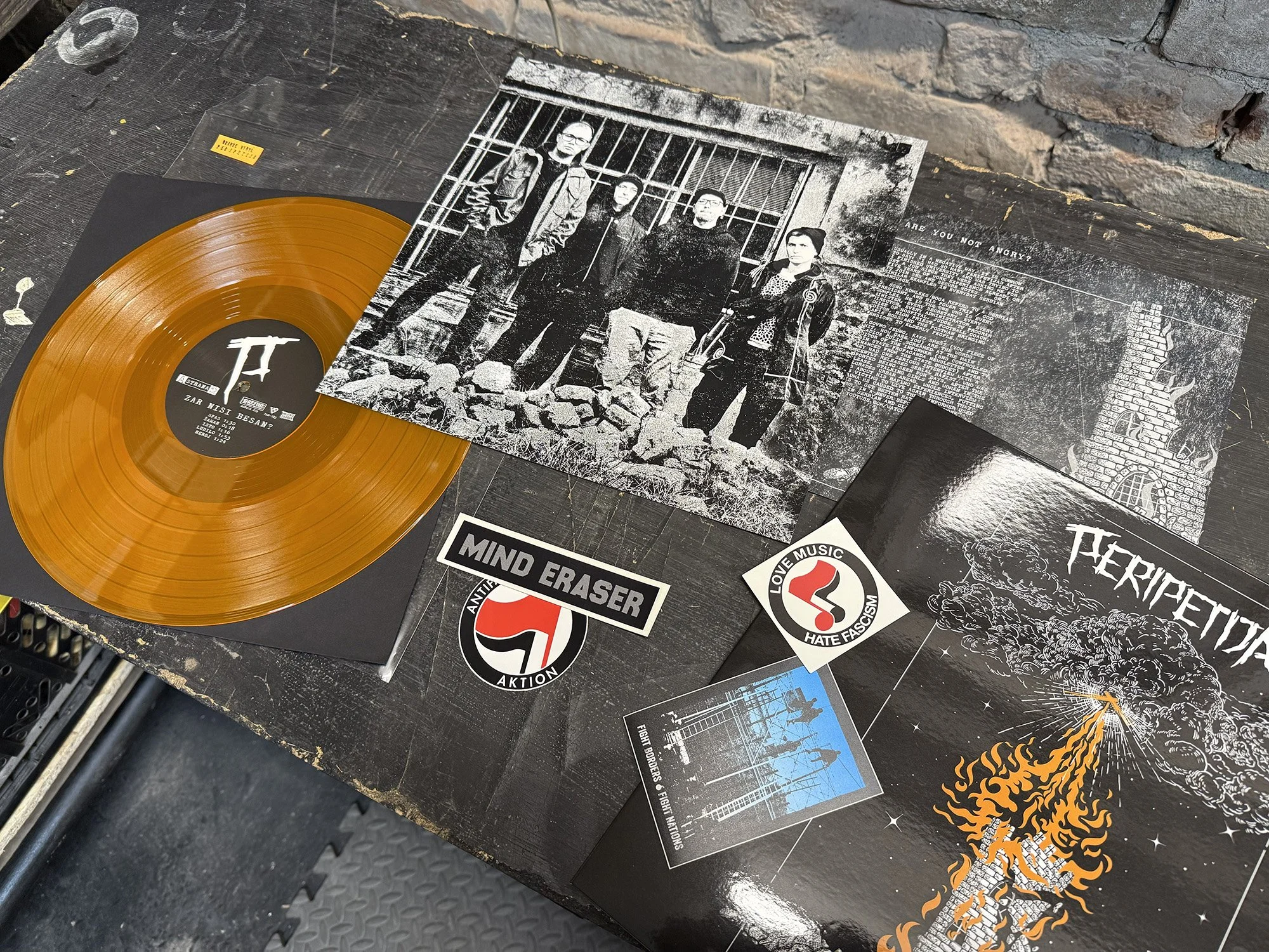 A vinyl record with a yellow-orange color and black center label resting on a black surface surrounded by posters, stickers, and artwork, including a black-and-white photo of four people standing in front of a barred gate, and stickers with anti-fasc