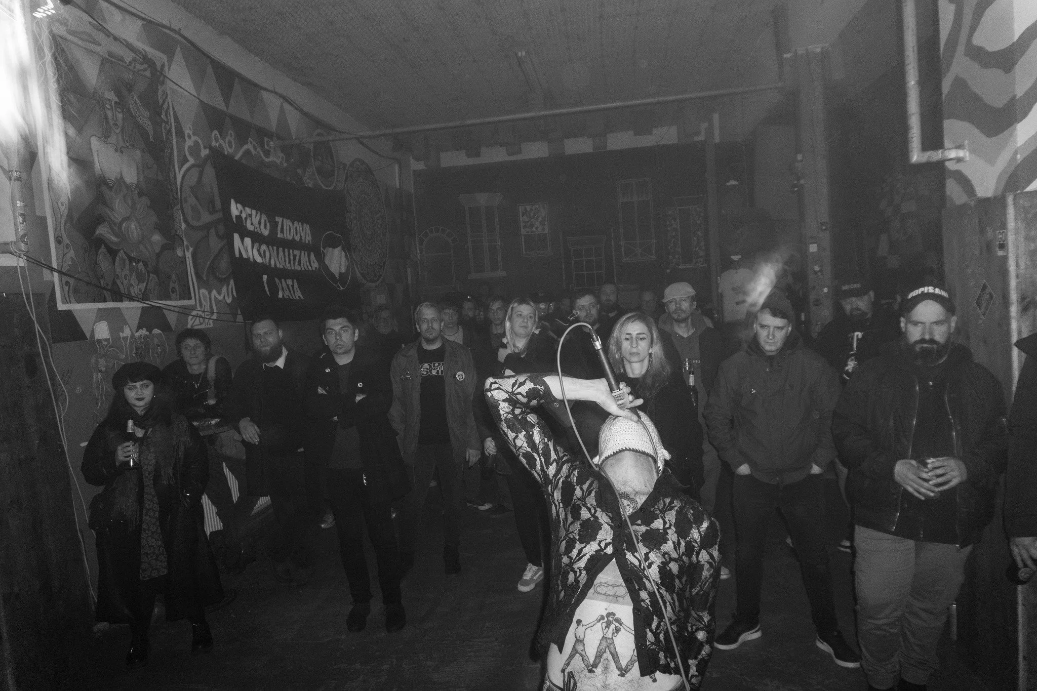 A group of people gathered indoors at an event, with a person in the foreground holding a microphone, and a banner with text in the background. The walls are decorated with colorful murals.