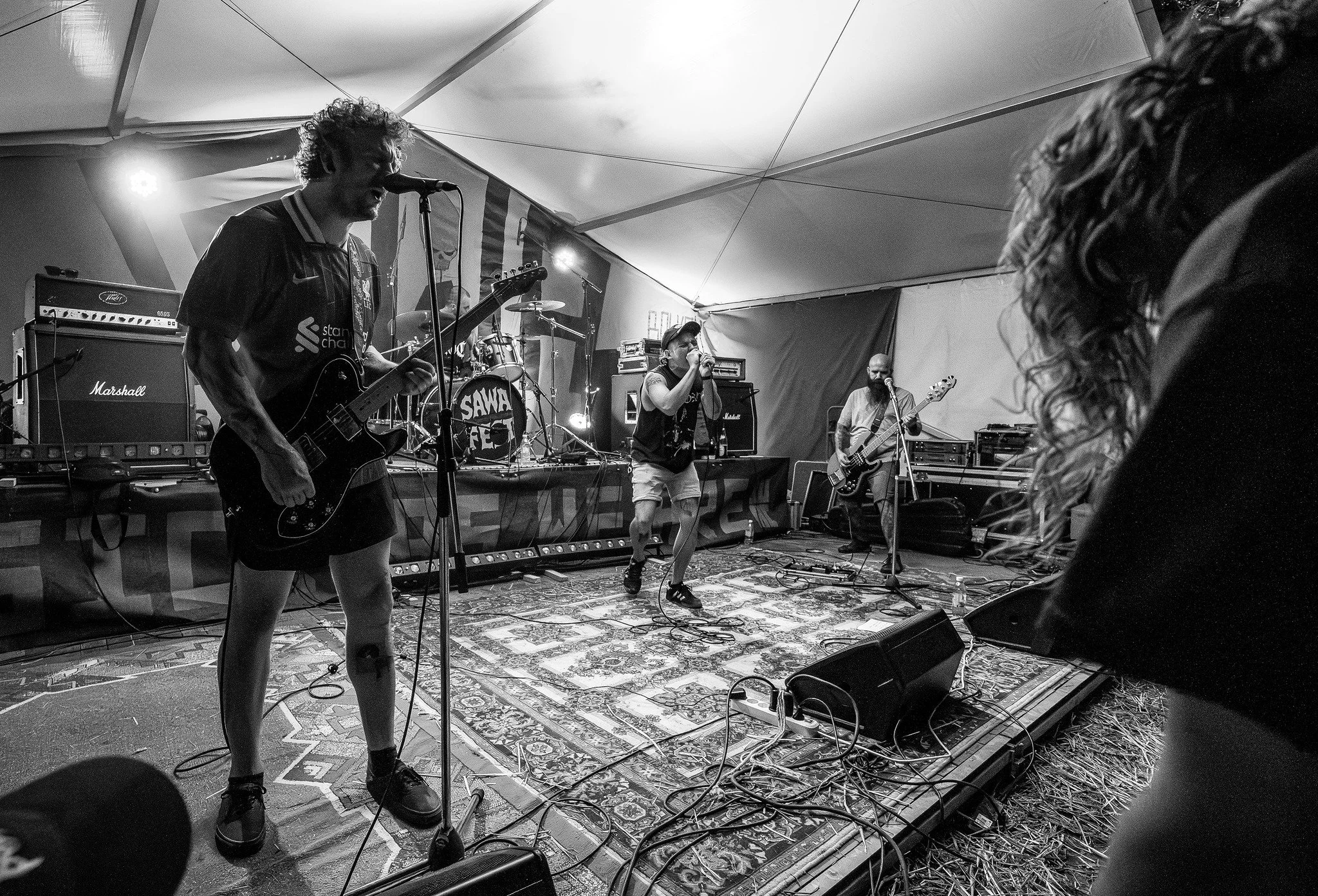 Black and white photo of a live music performance inside a tent. A guitarist and vocalist are in the foreground, with the guitarist playing an electric guitar. The vocalist is singing into a microphone. In the background, a drummer and a bassist are 