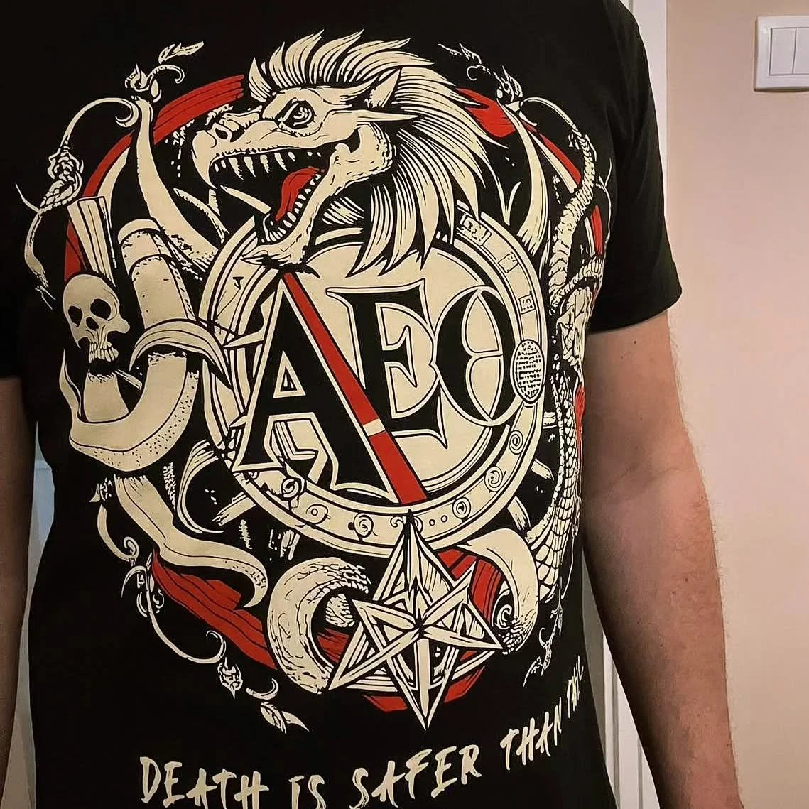 A black T-shirt with a graphic design featuring a fierce dragon, skull, snake, pentagram, and circular emblem with the letters 'AEO'. The T-shirt has white, red, and beige colors and the phrase 'Death is safer than' at the bottom.