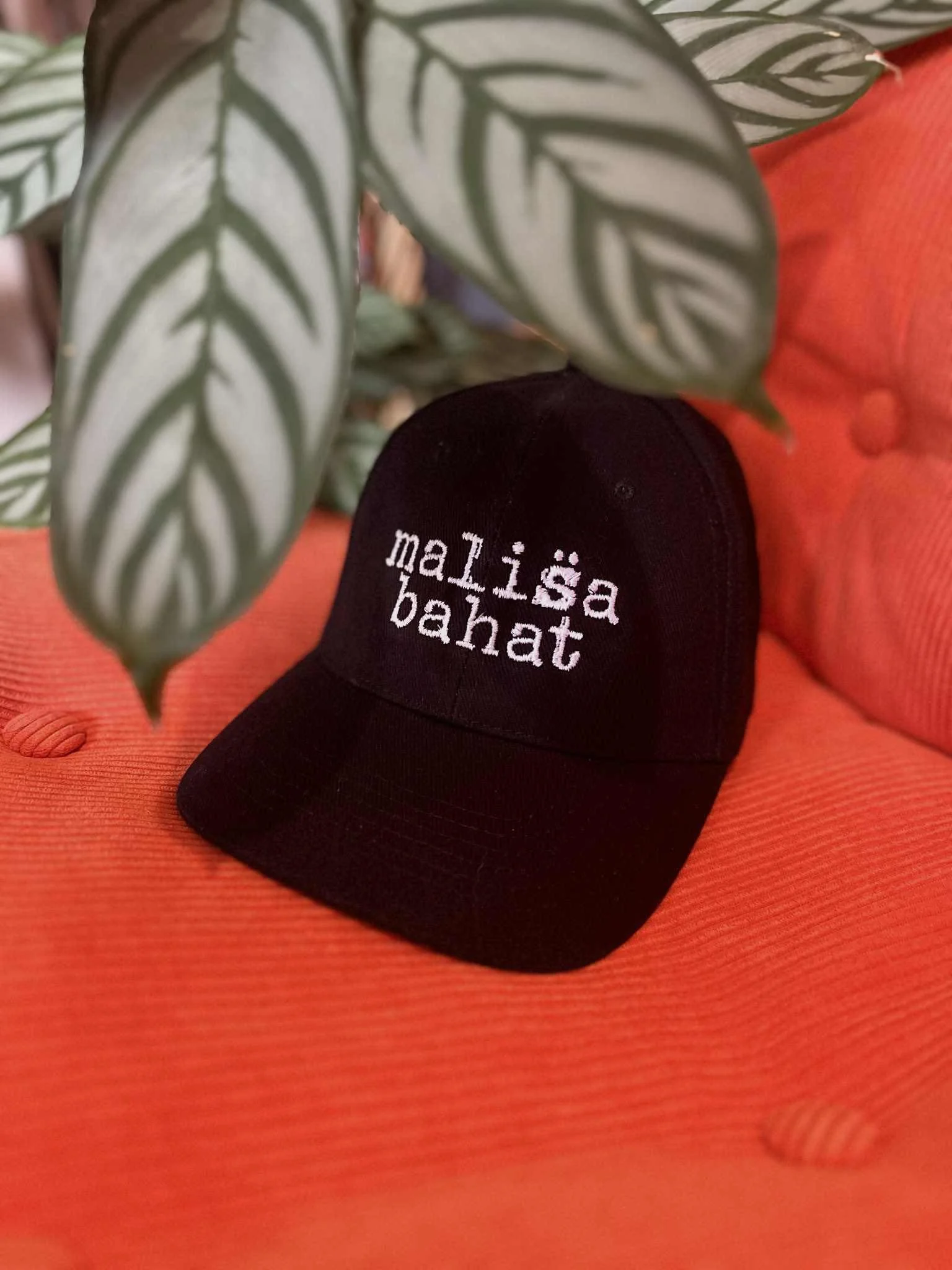 Black baseball cap with white embroidered text reading 'Maisha Bahat' on an orange textured surface.