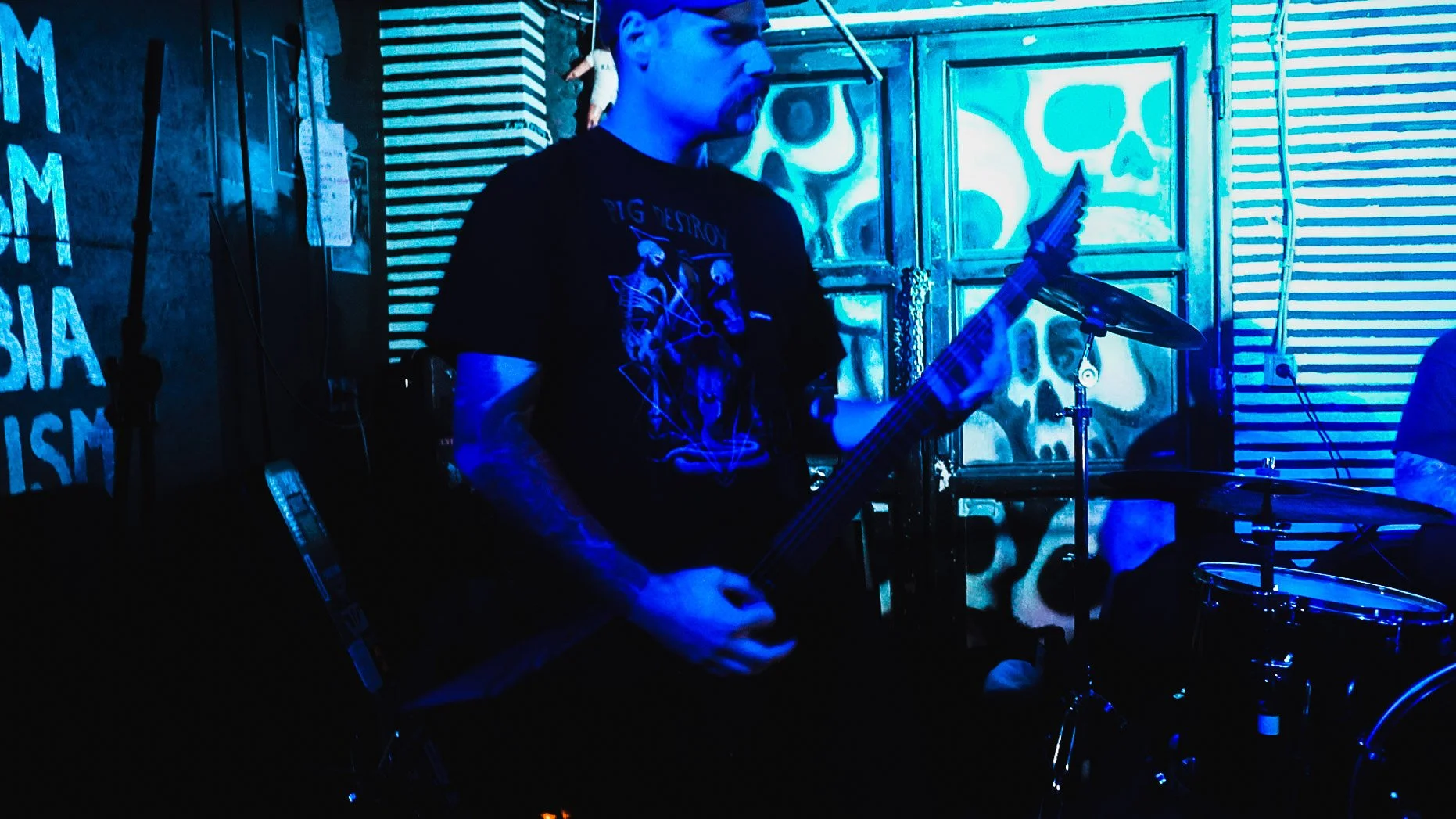 A man playing an electric guitar and a drum set in a dimly lit room with blue neon lighting, skull artwork on the background wall, and striped patterns on the wall to the right.