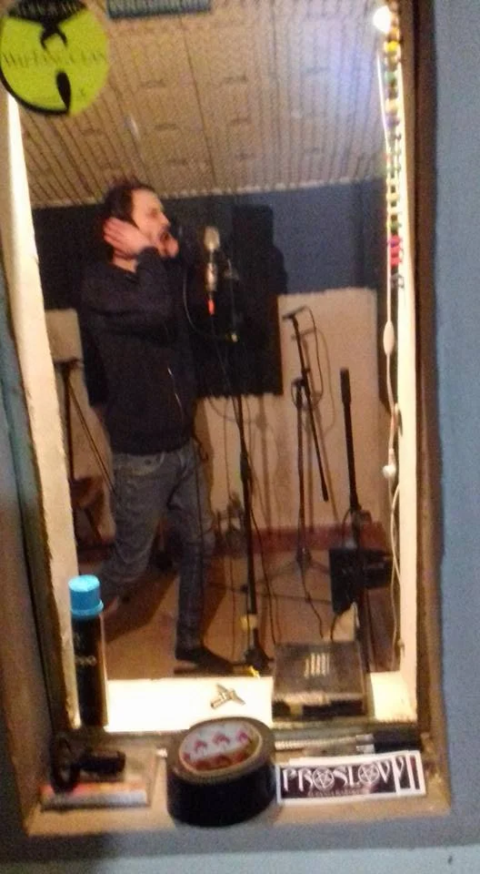 A man recording vocals in a music studio, standing in front of a microphone with headphones, in a room with soundproofing.