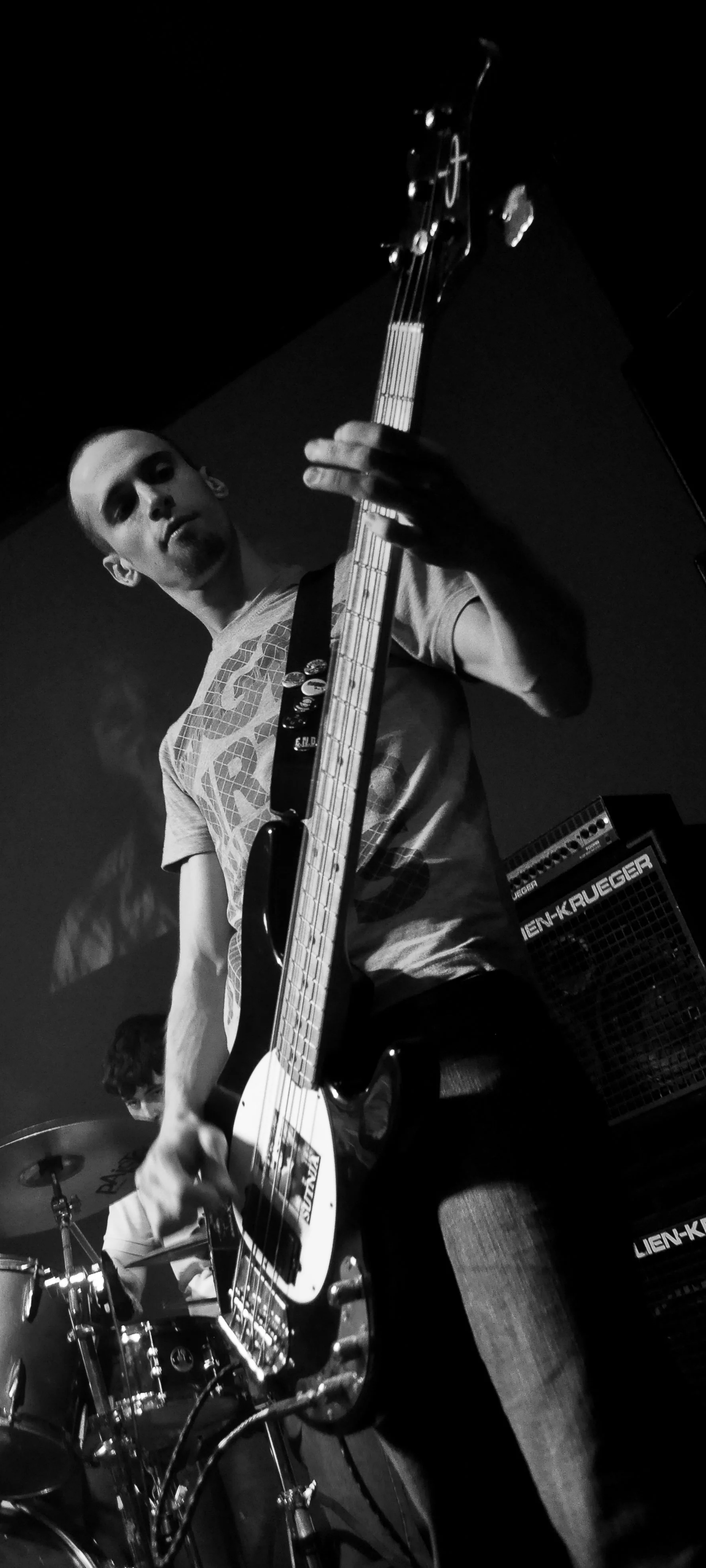 A young person playing an electric guitar on stage, with a drummer visible in the background, in black and white.