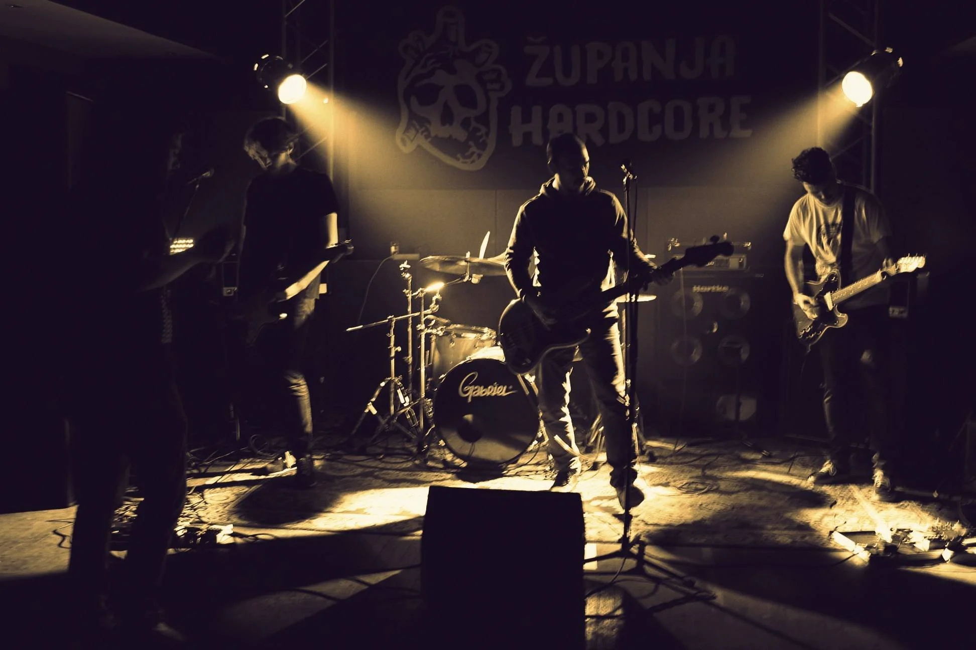 A band performing live on stage with four members playing guitar and drums, backlit by warm stage lighting, in front of a backdrop with a skull and the words 'Županja Hardcore'.