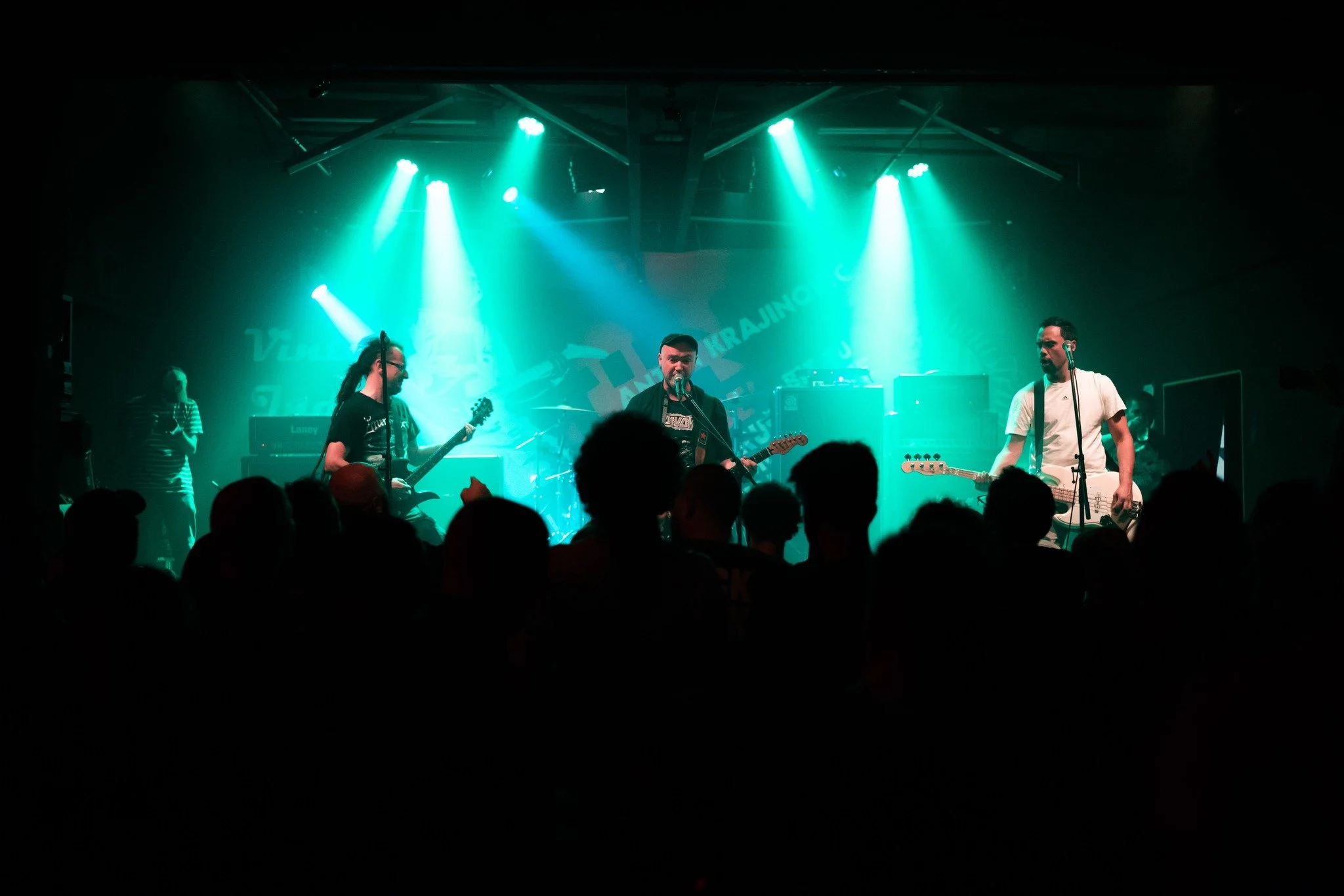 A band performing live on stage at a concert with blue-green stage lighting and audience in the foreground.