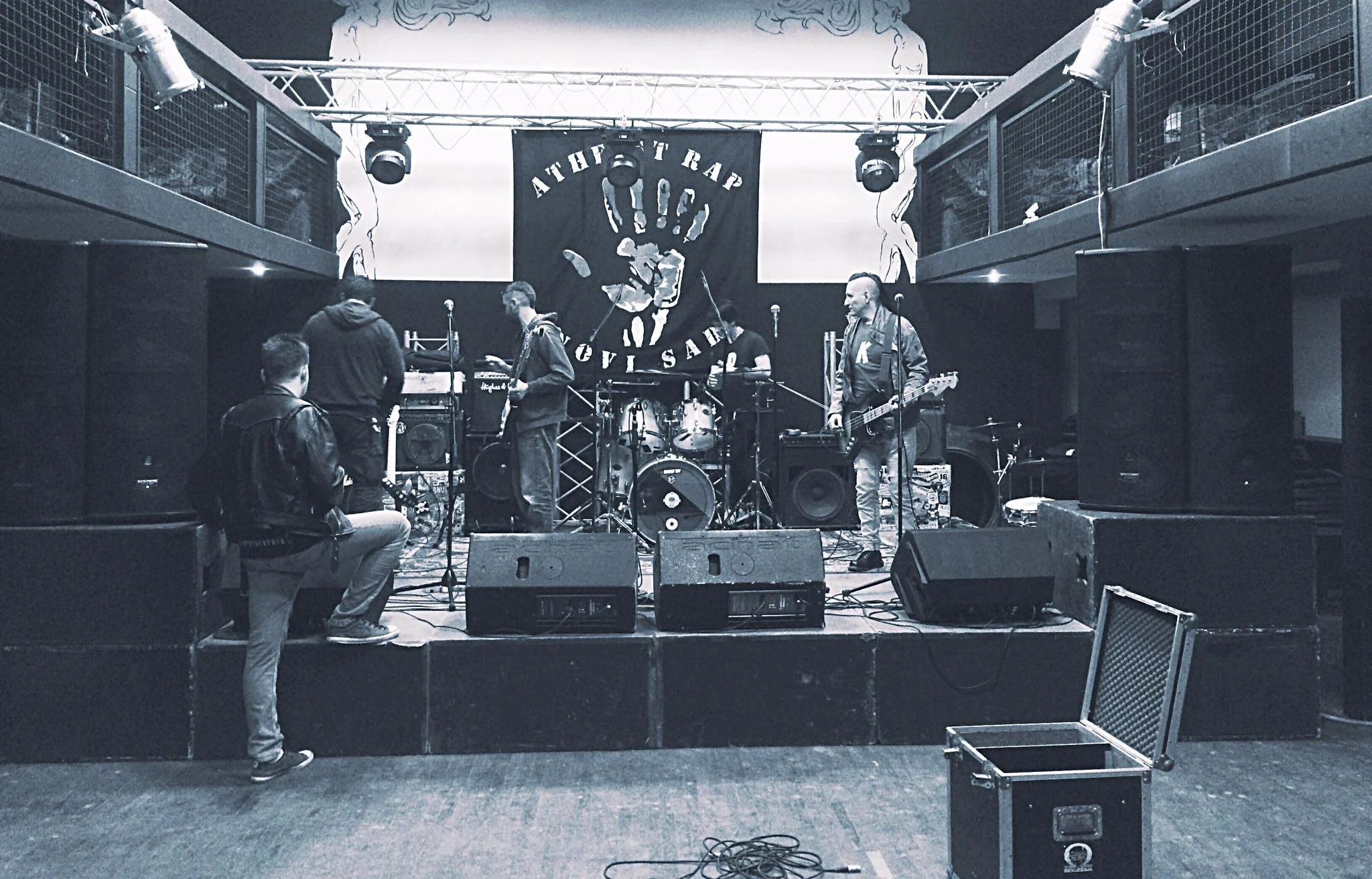 A music band setup on a stage with four musicians, including a drummer, two guitarists, and a bassist. The stage has lighting and speakers, with a large black banner hanging in the background featuring a hand and the words 'Atheist Rap'.