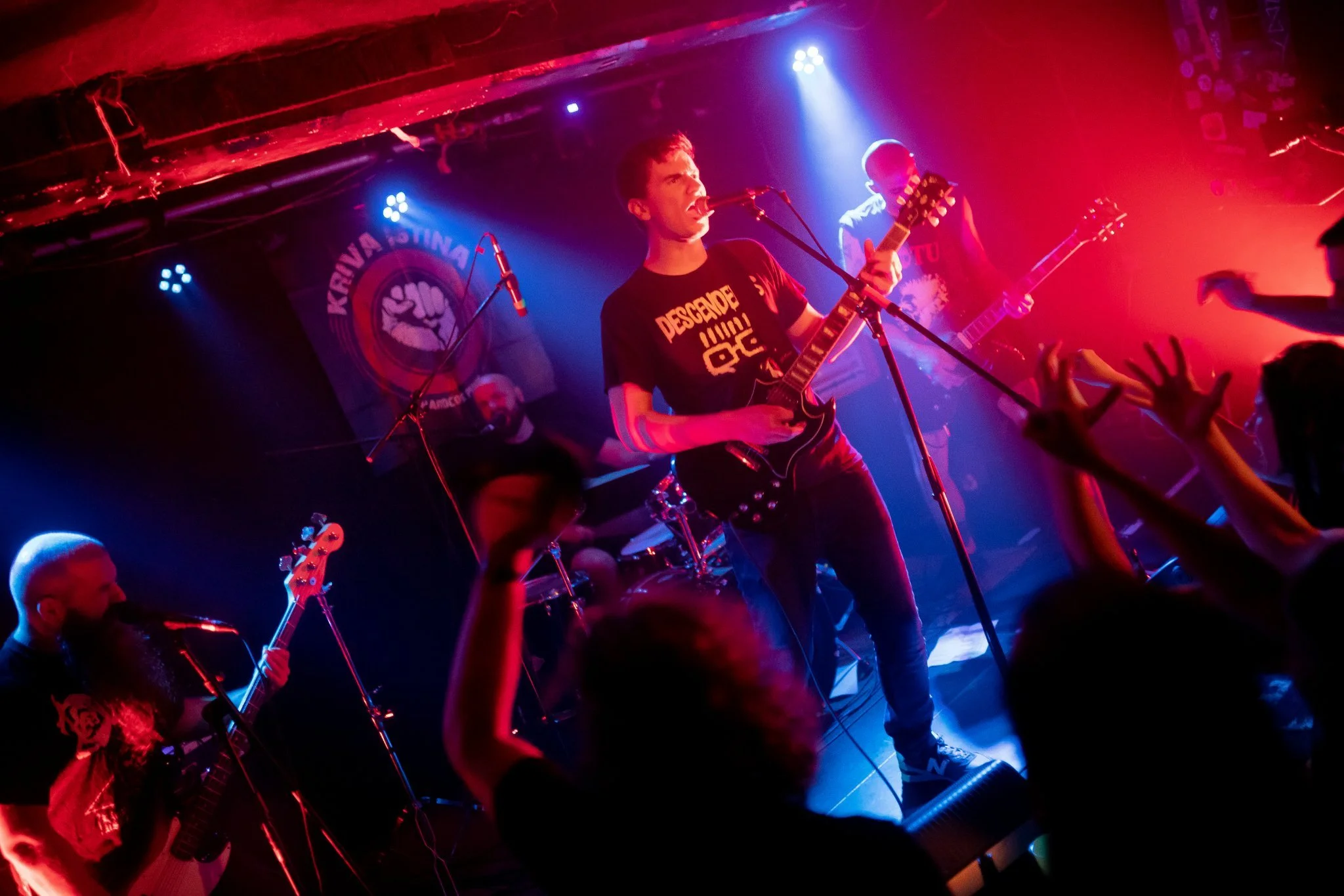 A band performs on stage with colorful lighting; the lead singer is playing an electric guitar and singing into a microphone, with a bassist and drummer visible in the background amidst concert audience.