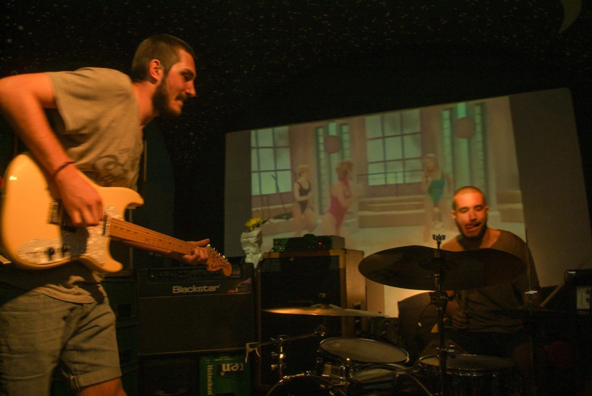 Two musicians performing indoors with a projection of animated characters on the wall behind them. One playing an electric guitar and the other on drums.
