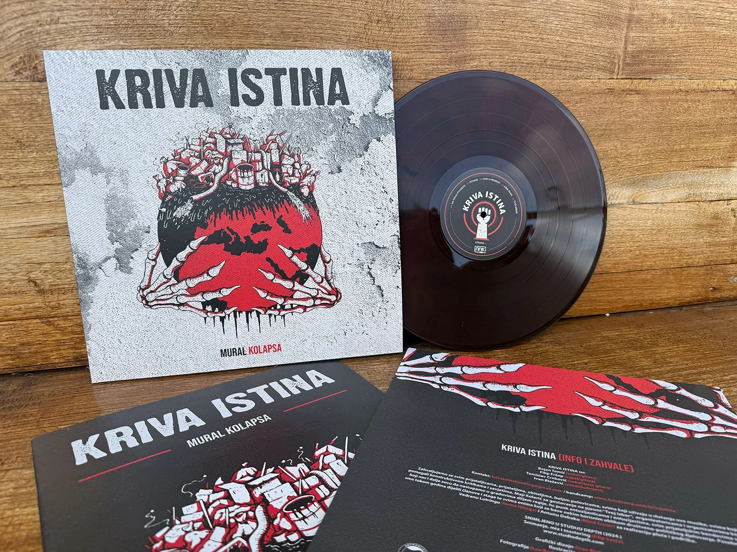 Vinyl record album titled 'KRIVA ISTINA' by Mural Kolapsa, with cover art featuring a red skull with hands and a crown of cans. The album is displayed on a wooden surface, with lyrics and promotional materials nearby.