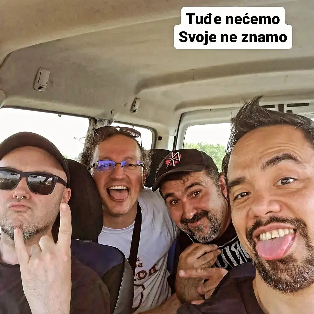 Four men taking a selfie inside a vehicle. One man is wearing sunglasses and a septum piercing, making a rock sign. The others are smiling and making various hand gestures. A text overlay in a foreign language is present at the top of the image.