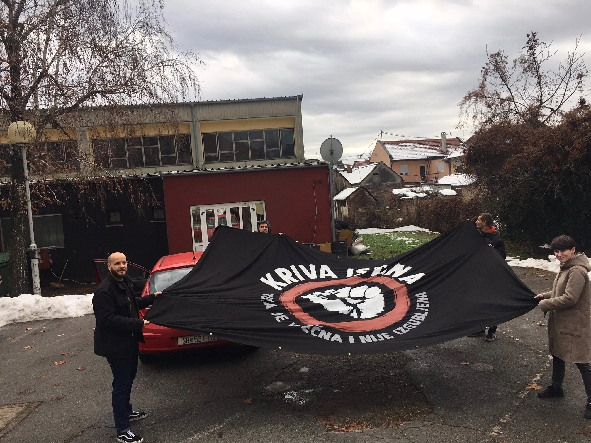 A group of people holding a large black flag with white and red text and a red circle symbol. The scene is outdoors on a cloudy winter day with snow on the ground, and trees and buildings in the background.