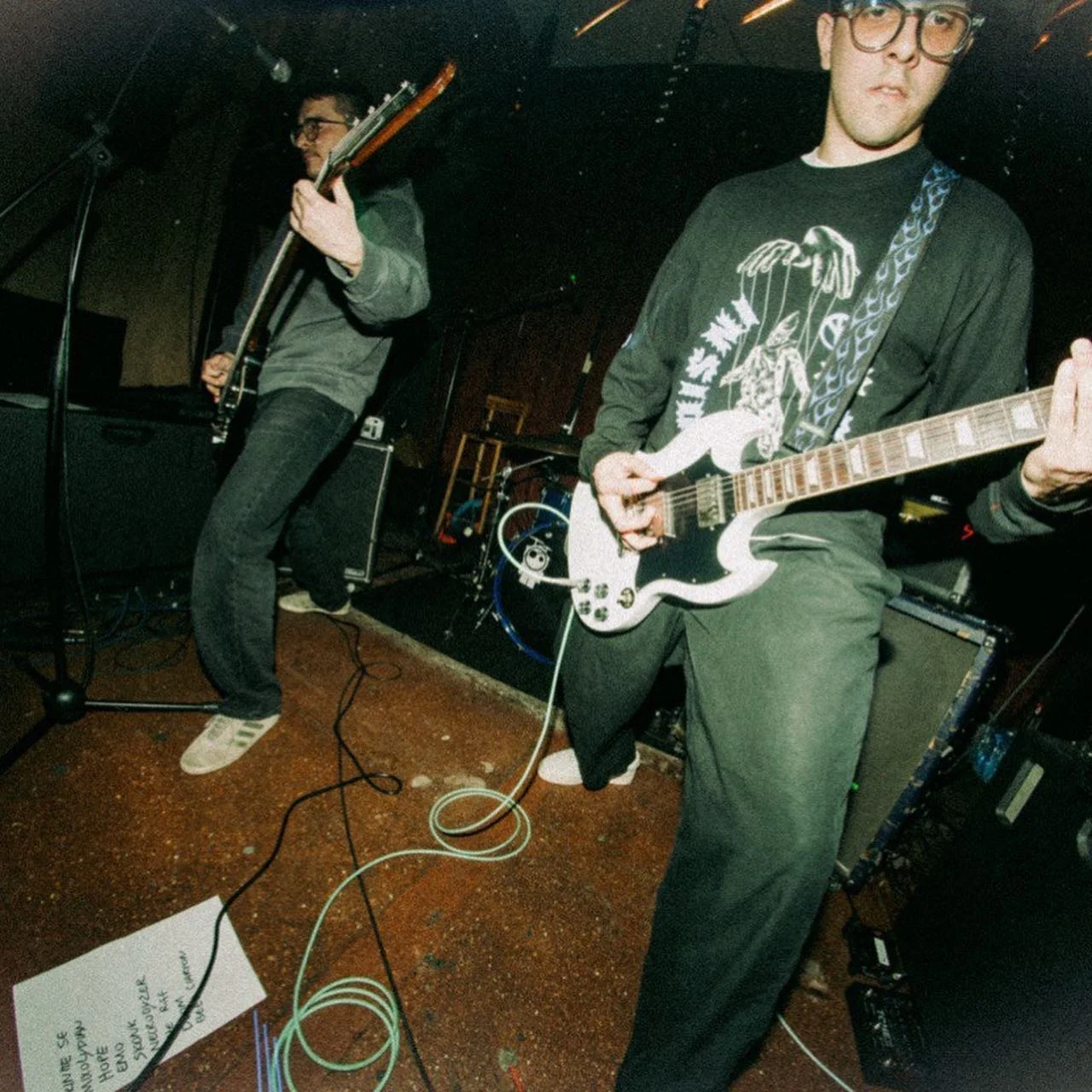 Two musicians performing on stage, one playing an electric guitar and the other playing a bass. There are various musical equipment and cords on the floor, with a sheet of paper nearby.