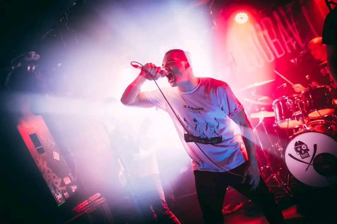 A vocalist passionately singing into a microphone during a live rock concert with red and purple stage lighting, a drummer with skull and crossbones brand on drum kit in the background.
