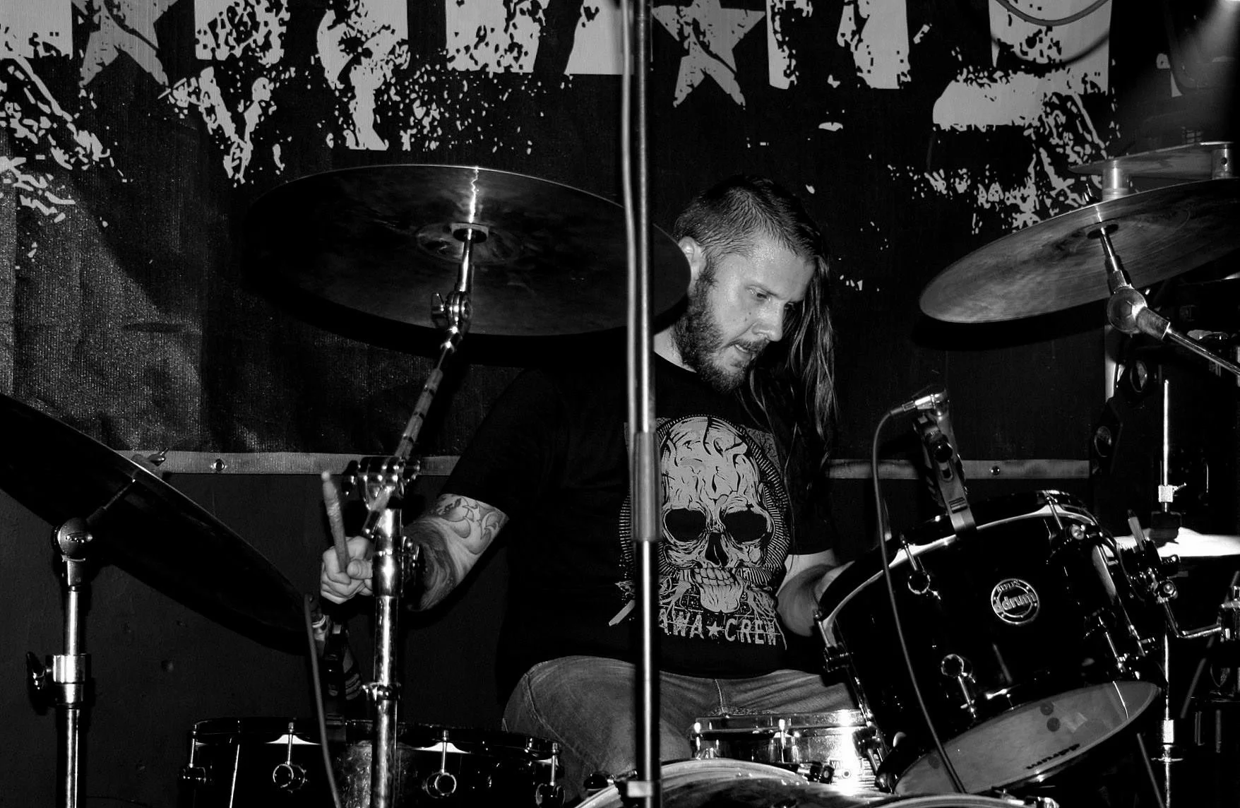 A man with long hair and a beard playing drums in a black and white setting, wearing a T-shirt with a skull graphic, in a dimly lit room with a textured backdrop.