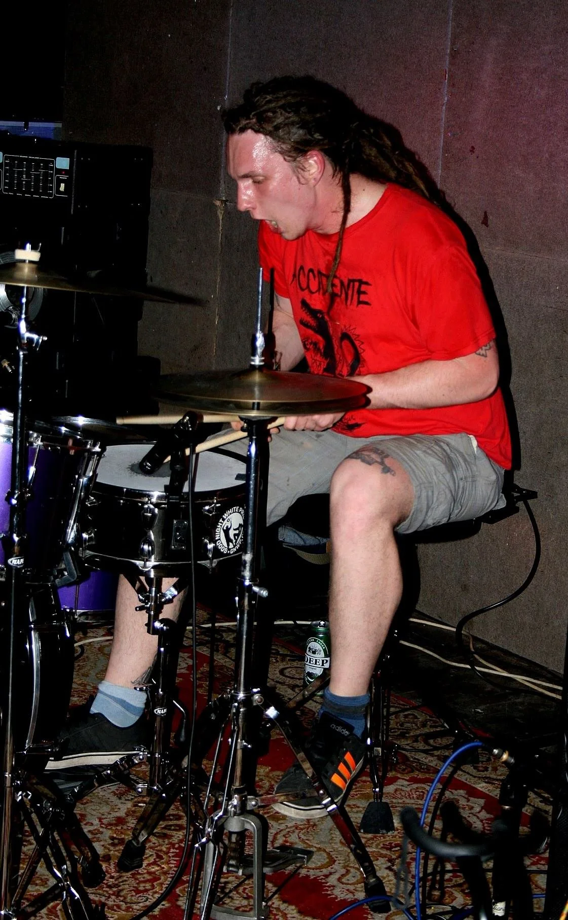 A man with long dreadlocks wearing a red T-shirt and grey shorts is playing the drums. He appears focused and passionate, sitting on a drum throne with a small patterned rug underneath. There is a soft drink can on the floor near his foot. The backgr