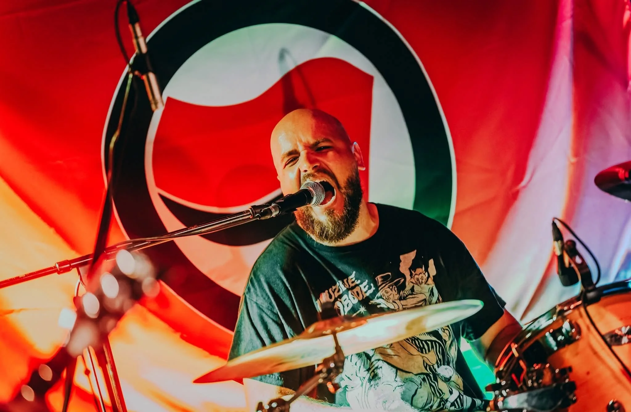 A man with a beard and a shaved head singing into a microphone while playing drums during a performance, with a large antifa logo banner in the background, colorful stage lighting.