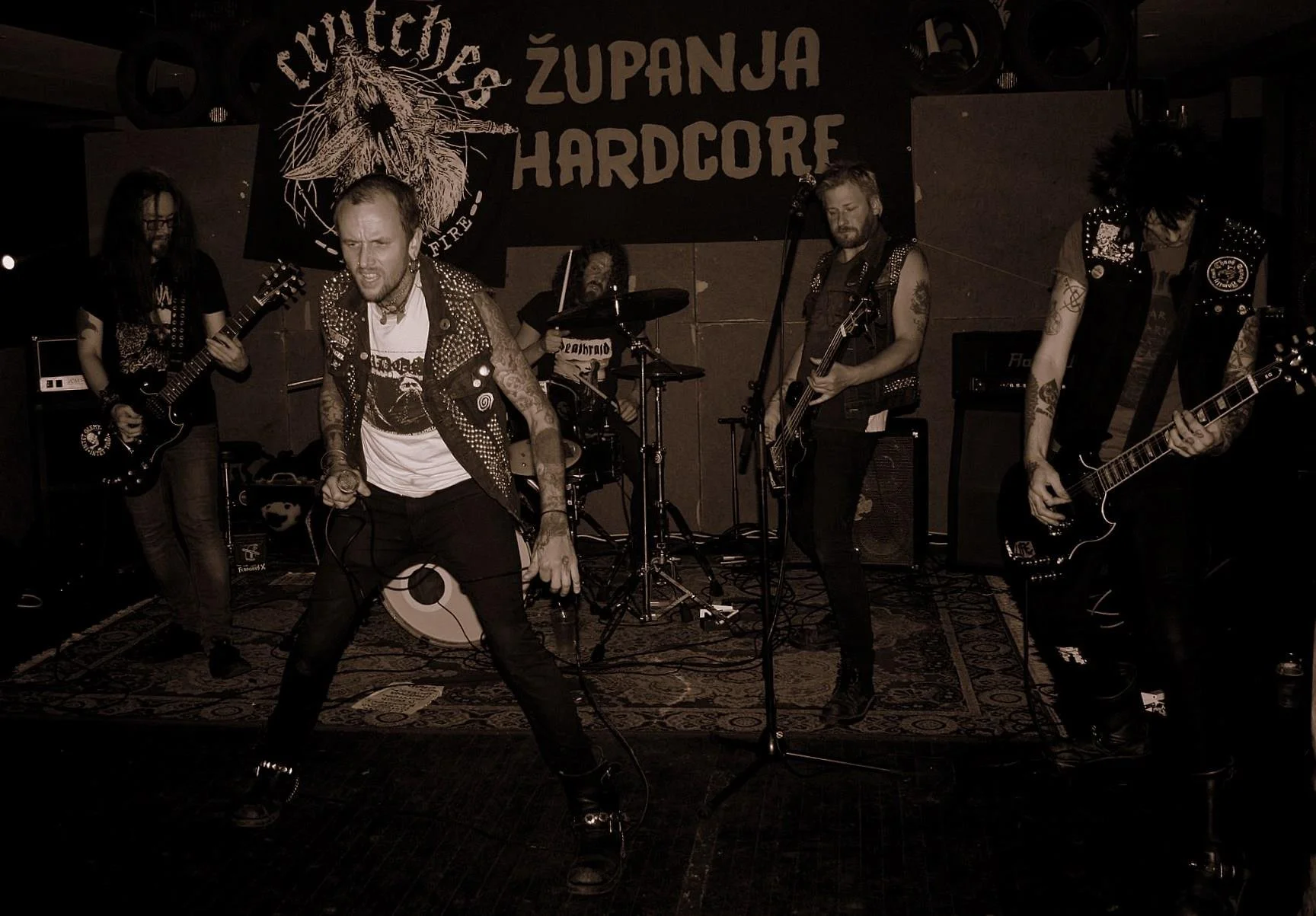 A band performs live on stage with five members, including a lead singer with tattoos and spiked vest, two guitarists, a bassist, and a drummer. A large banner in the background reads "ŽUPANJA HARDCORE" with a wolf graphic.