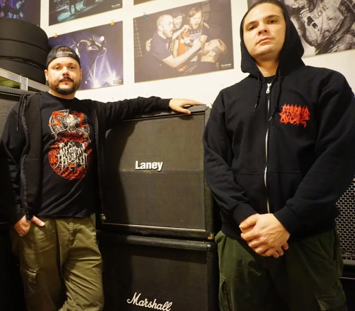 Two men standing next to music equipment in a room with posters on the wall. The man on the left wears a black cap backward, a black hoodie with red graphic, and khaki pants. The man on the right wears a black hooded jacket and green pants. The equip