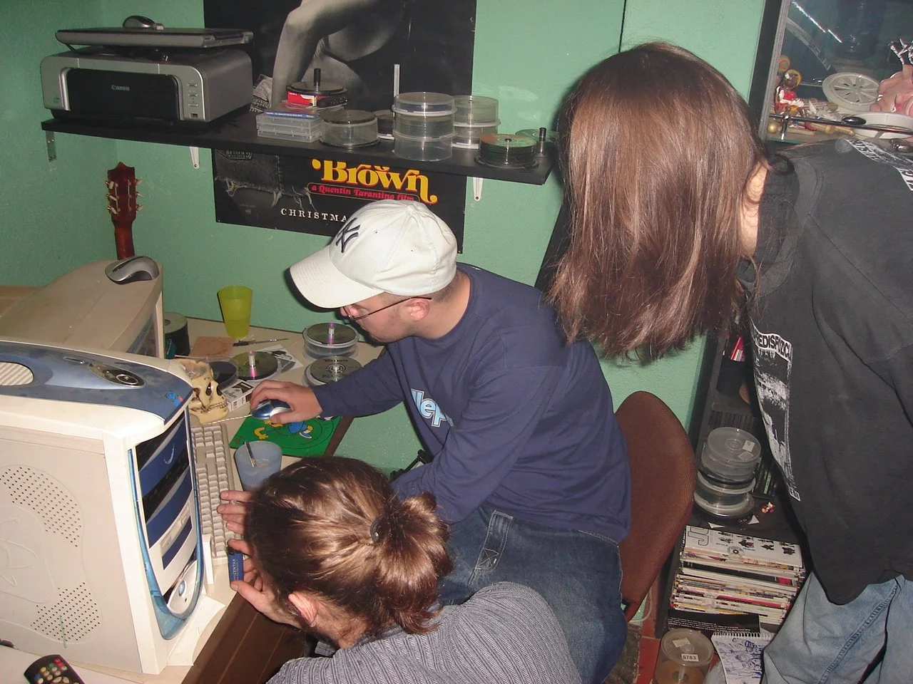 Three young people gathered around an old computer monitor in a room filled with CDs, film reels, and posters. One person is seated at the computer wearing a gray sweater, a person with glasses and a white cap is working on the computer, and the thir