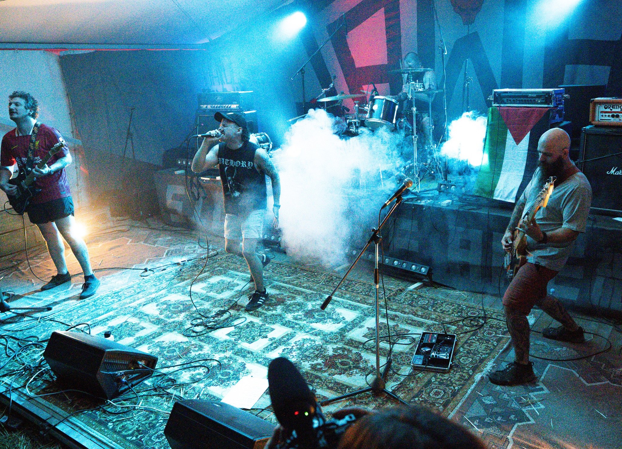 A band performing live on stage with musical instruments including a guitar, drums, and microphone, with colorful lighting and smoky effects in the background.