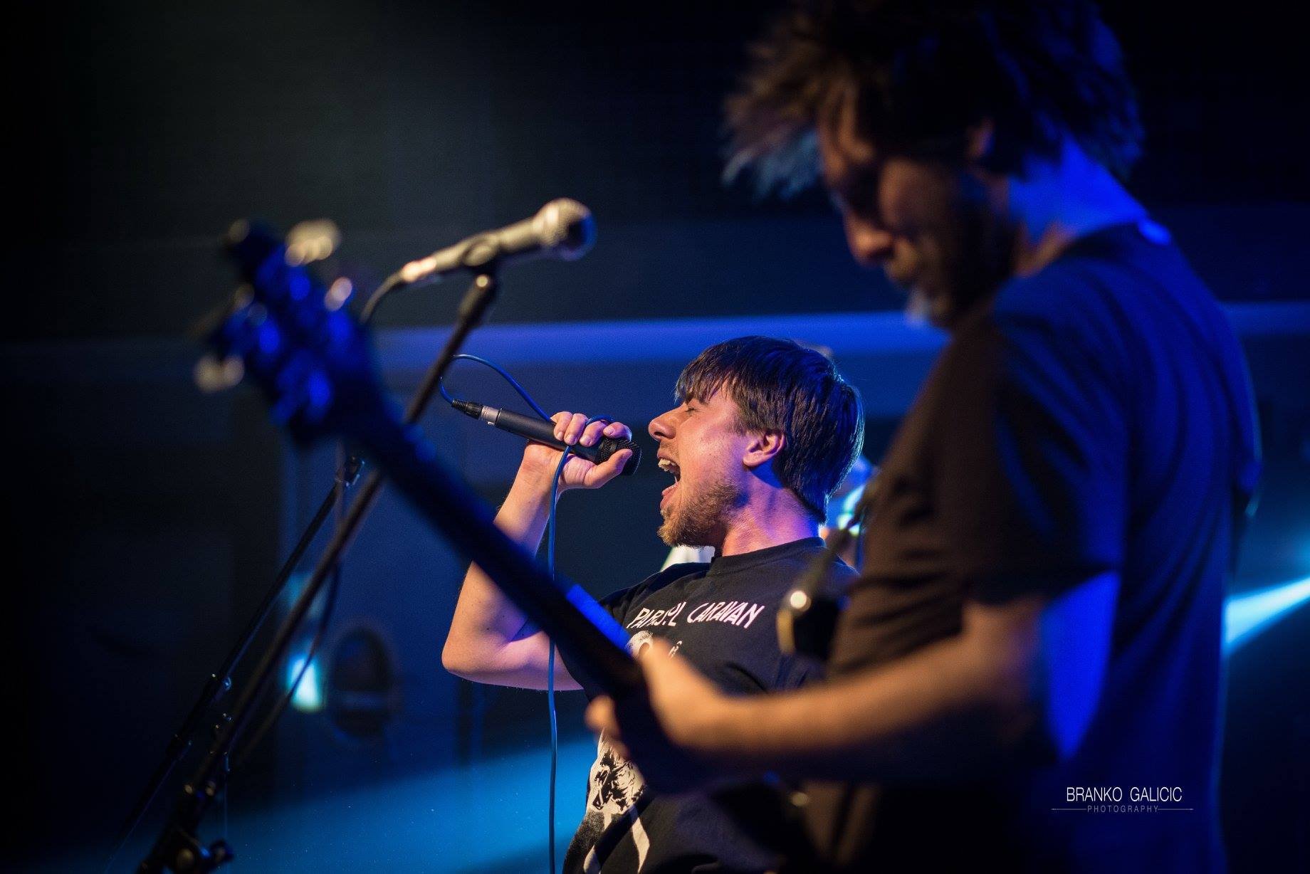 Two musicians perform on stage under blue lighting; one is singing into a microphone with closed eyes, and the other is playing guitar with a focused expression.