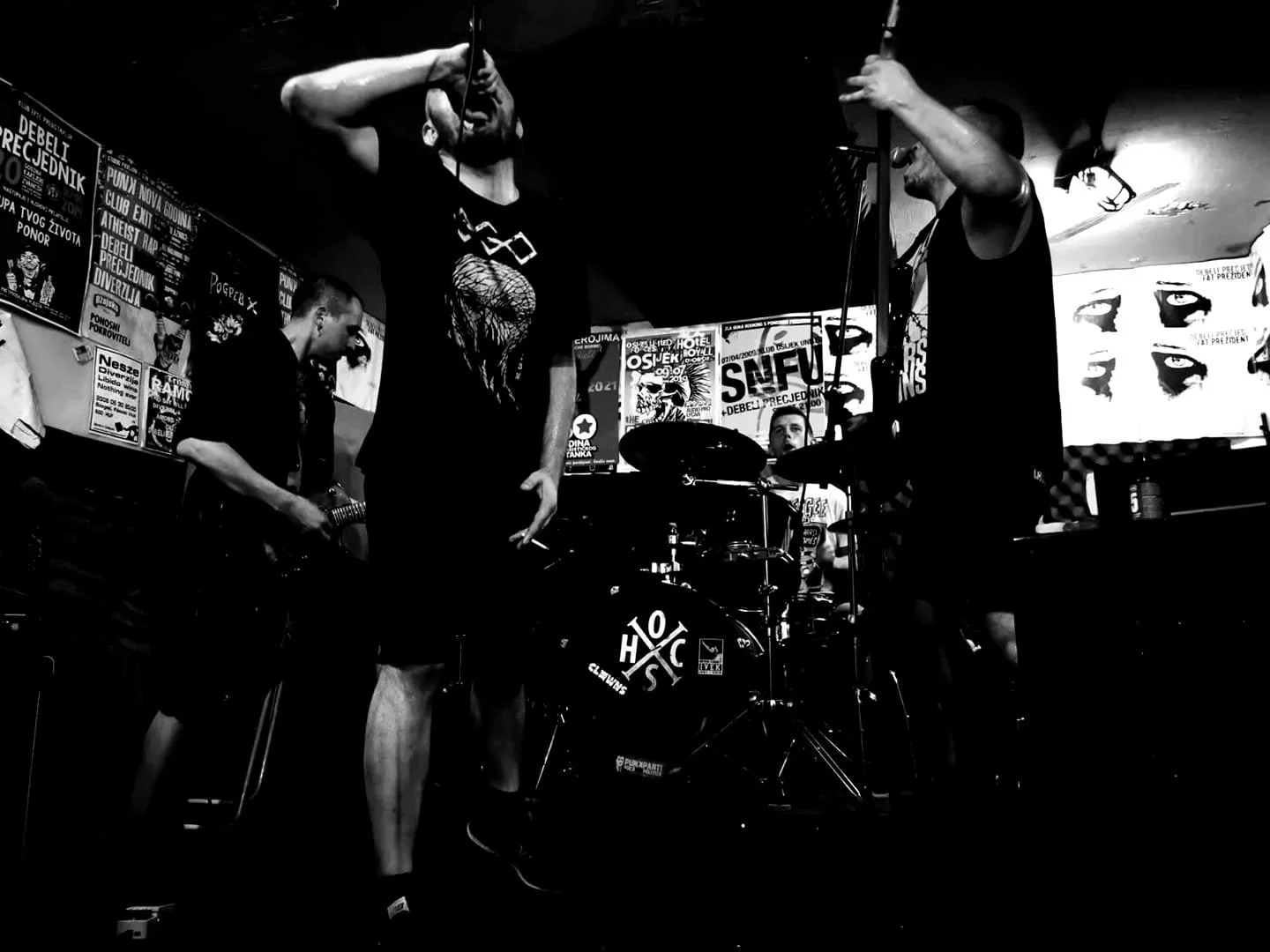 Black and white photo of a band performing on stage with three musicians; one singing into a microphone, one playing electric guitar, and one on drums. Posters on the wall are visible in the background.