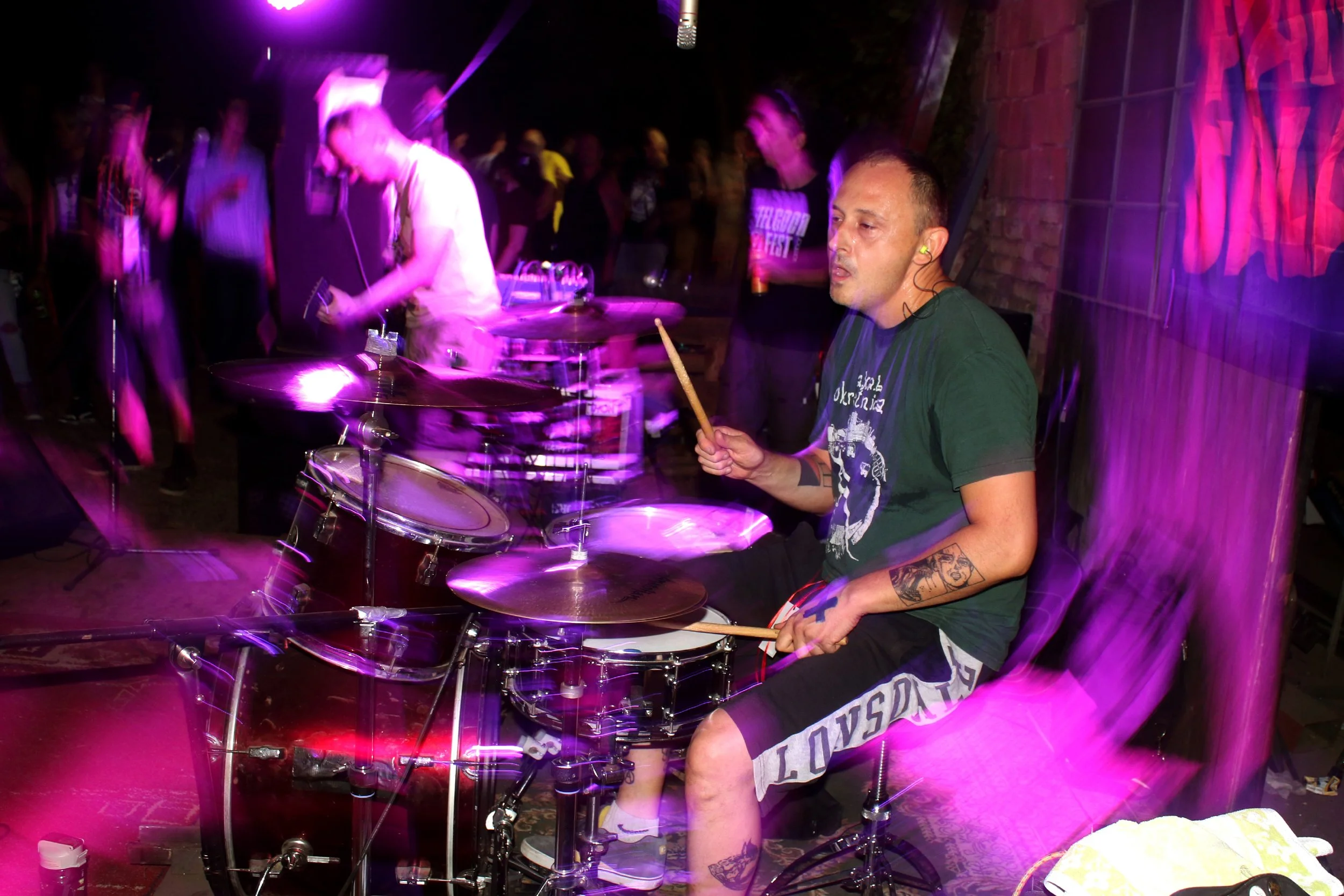 A man playing drums in a dimly lit music venue with purple lighting, surrounded by other people enjoying the performance.
