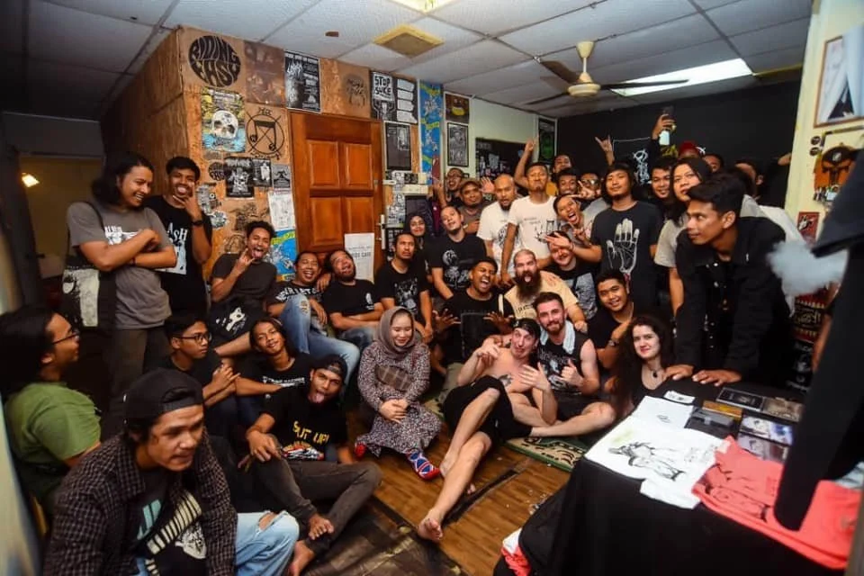 Group of diverse young people gathered in a cozy art studio or cafe, smiling and posing for a photo, with art posters and artworks on the walls.