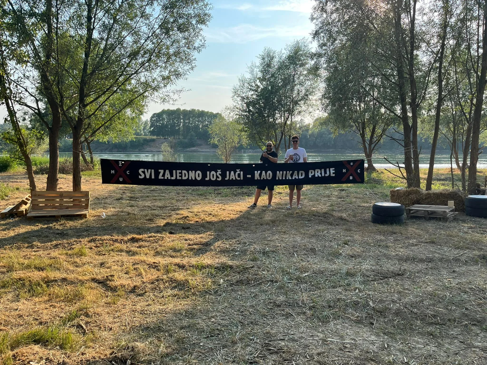 Two men standing behind a long banner that reads 'SVI ZAJEDNO JOŠ JAČI - KAO NIKAD PRIJE,' with trees and a river in the background on a sunny day.