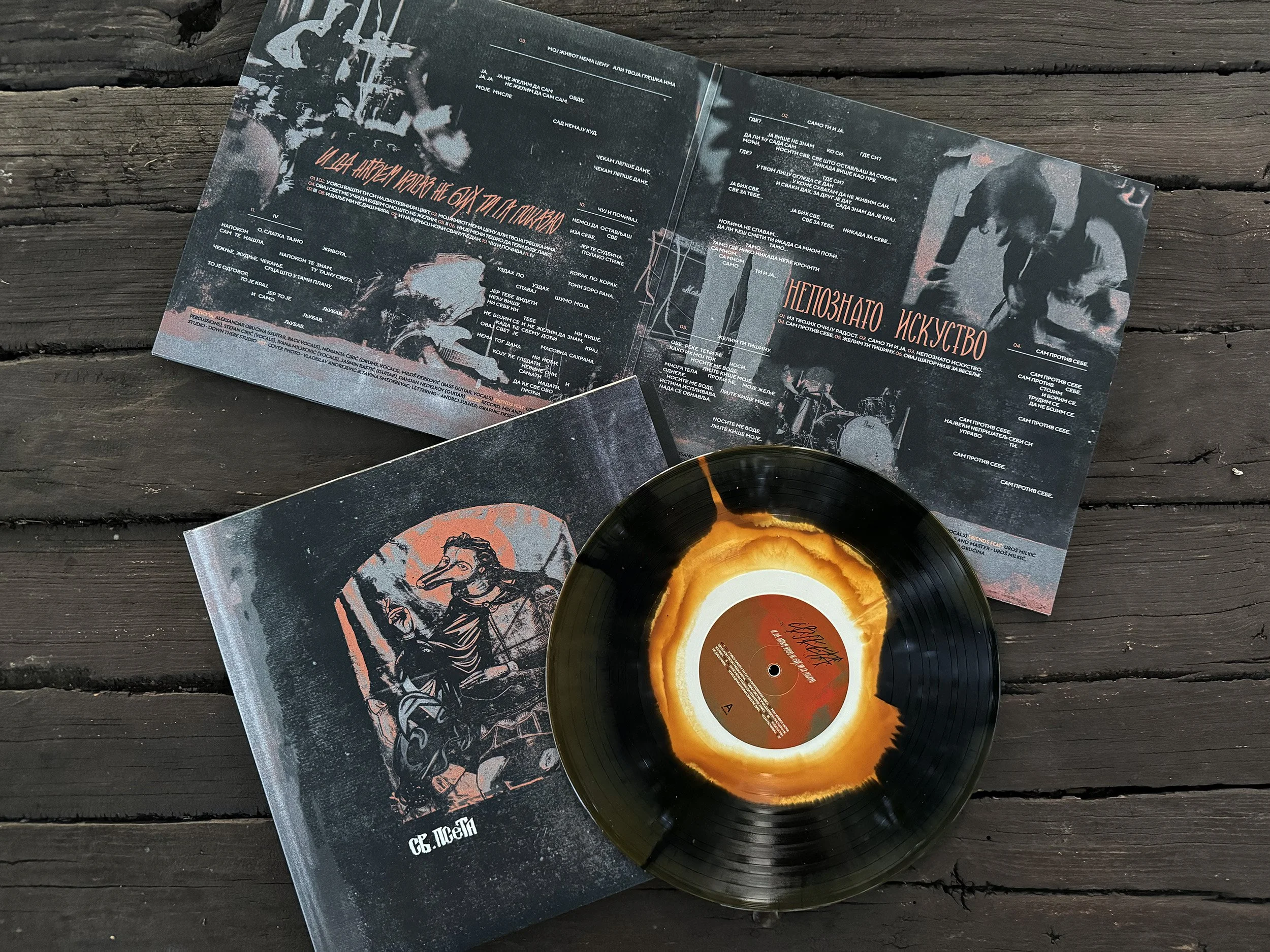 A vinyl record with a black and orange swirl pattern resting on a dark wooden surface. Behind the record, there are open booklets or magazines with artistic and textual content, including illustrations of people and abstract art. SVETA PSETA