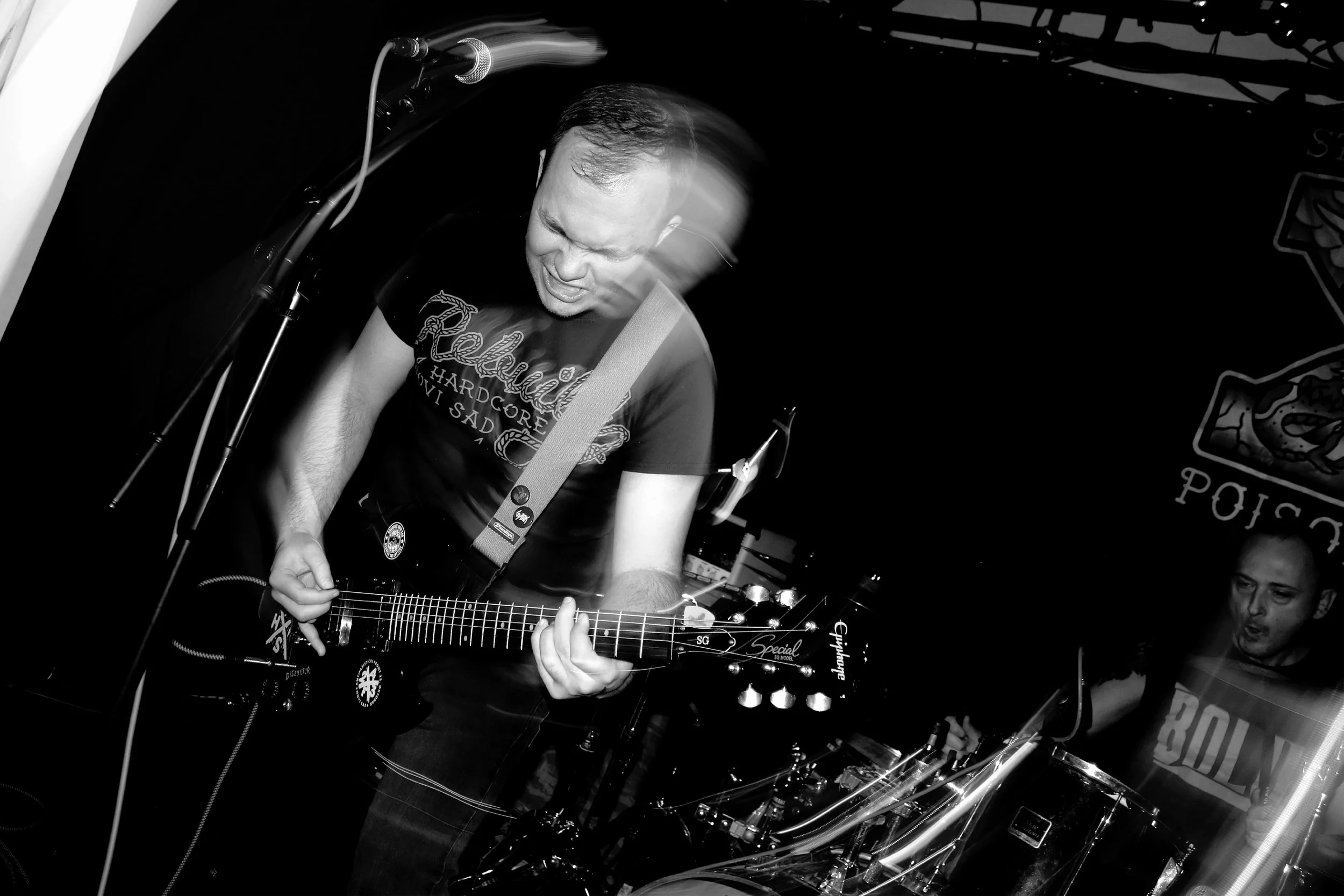 Black and white photograph of a man passionately singing and playing an electric guitar on stage, with motion blur and a microphone overhead.