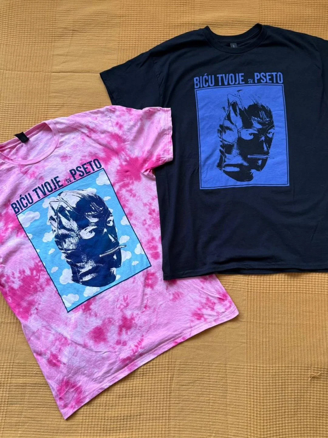 Two T-shirts, one pink tie-dye and one black, lying on a wooden surface, both featuring the same graphic design of a stylized dog's face with text in a foreign language