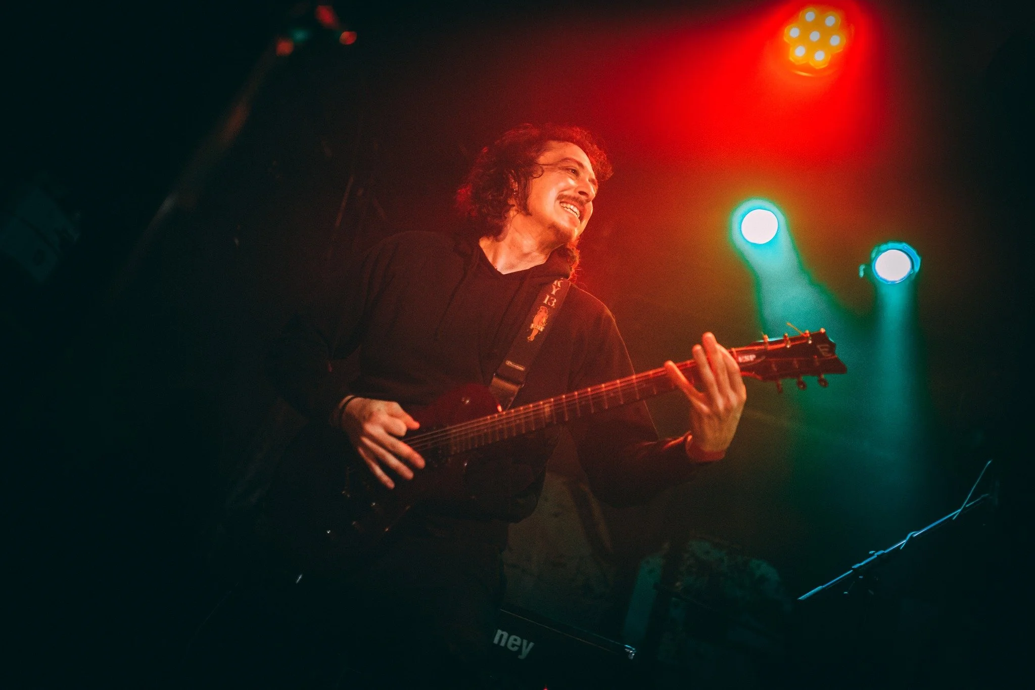A man with long curly hair playing an electric guitar on stage, illuminated by colorful stage lights.
