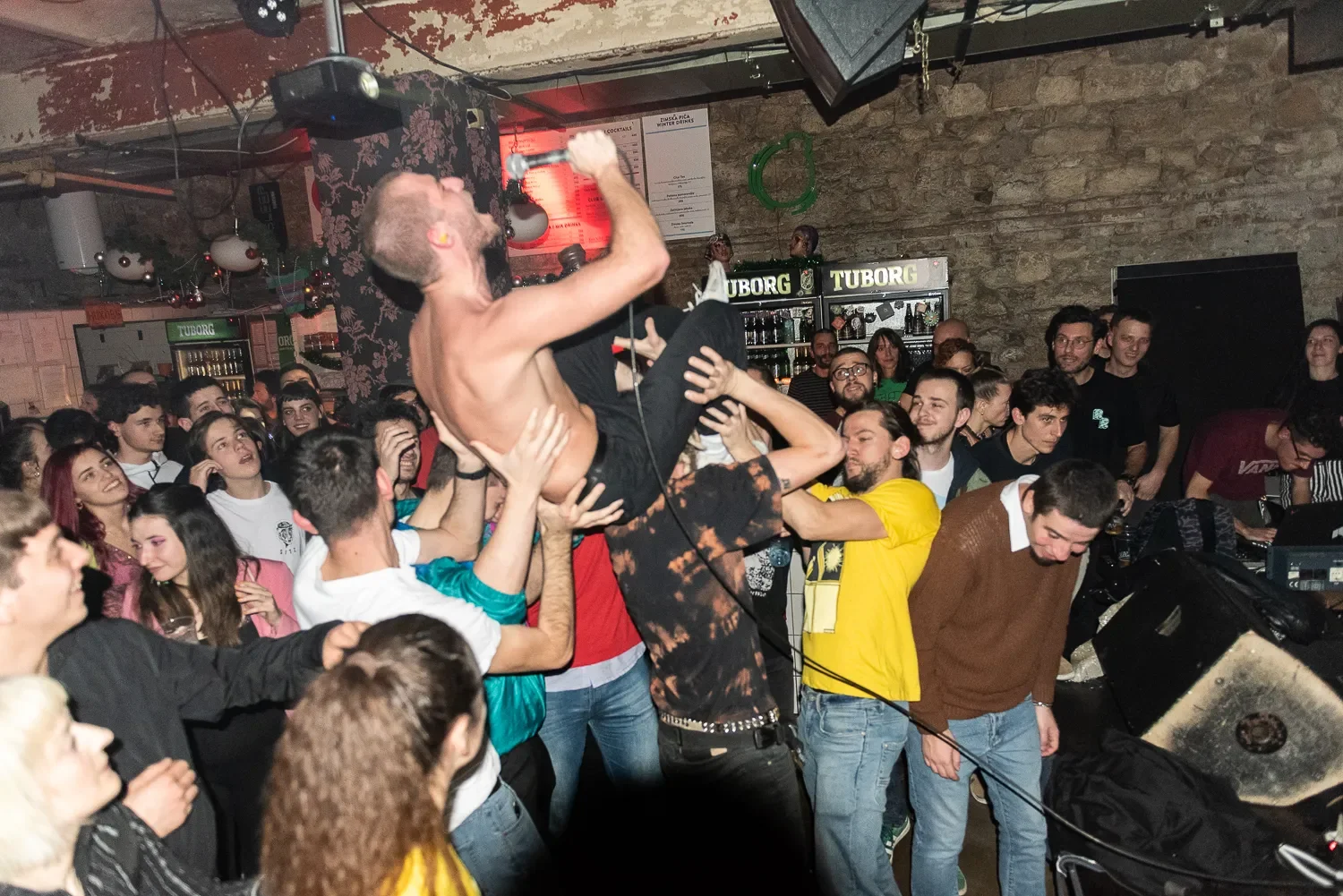 A shirtless male performer being lifted onto the crowd at a concert or party. The crowd is lively and diverse, with some people reaching out to support him. The background shows a bar area with signs for Tuborg beer and a stone wall.